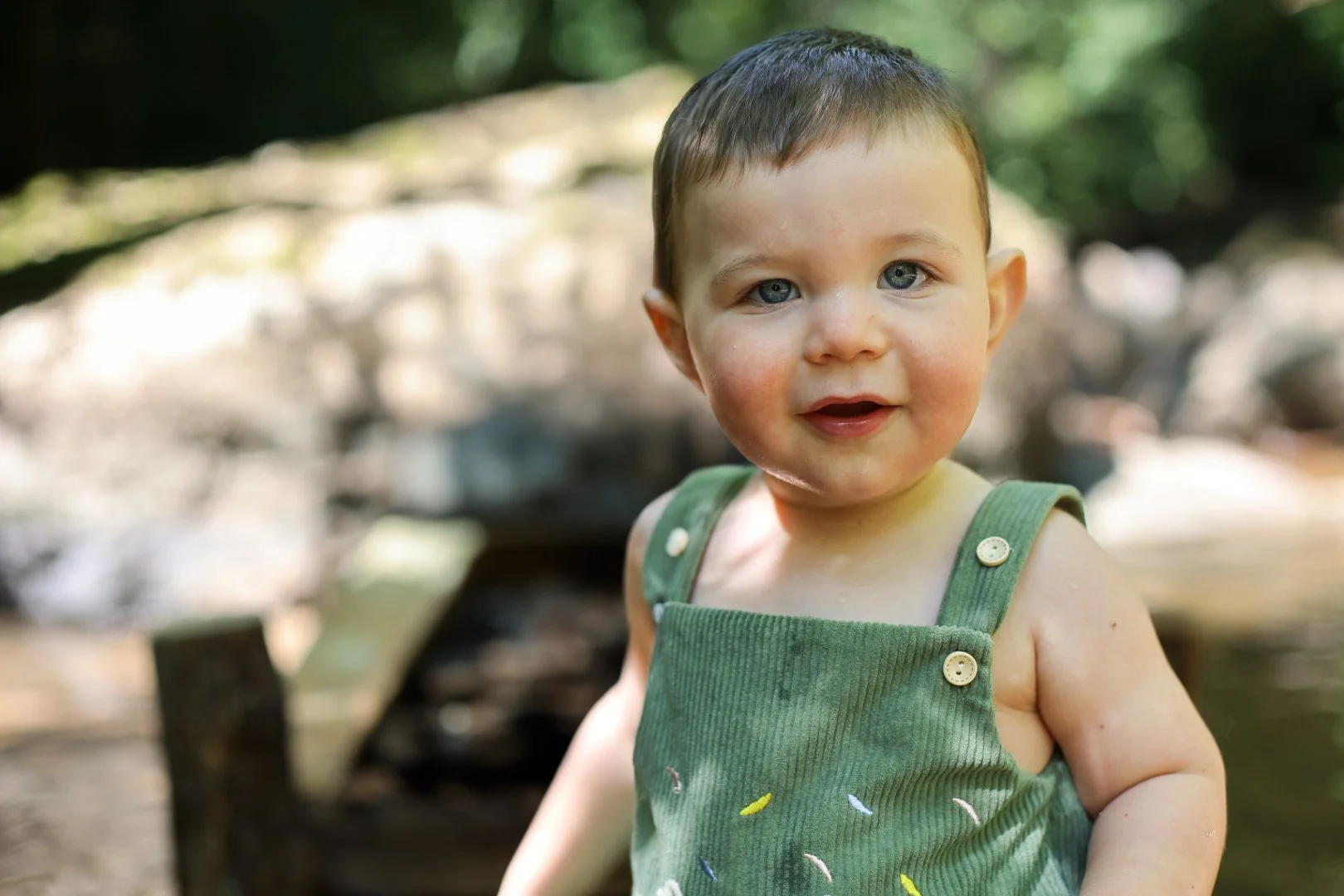 Un jeune enfant avec des cheveux courts, portant un ensemble vert à boutons, se trouve dans un environnement naturel avec des arbres et de la lumière filtrée.