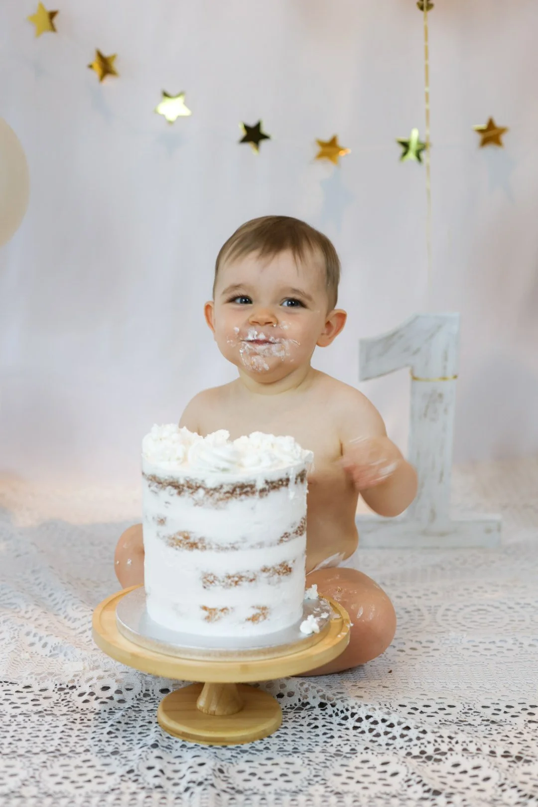 Un bébé souriant assis sur une nappe en dentelle avec un gâteau à la crème devant lui, lors d'une fête d'anniversaire.