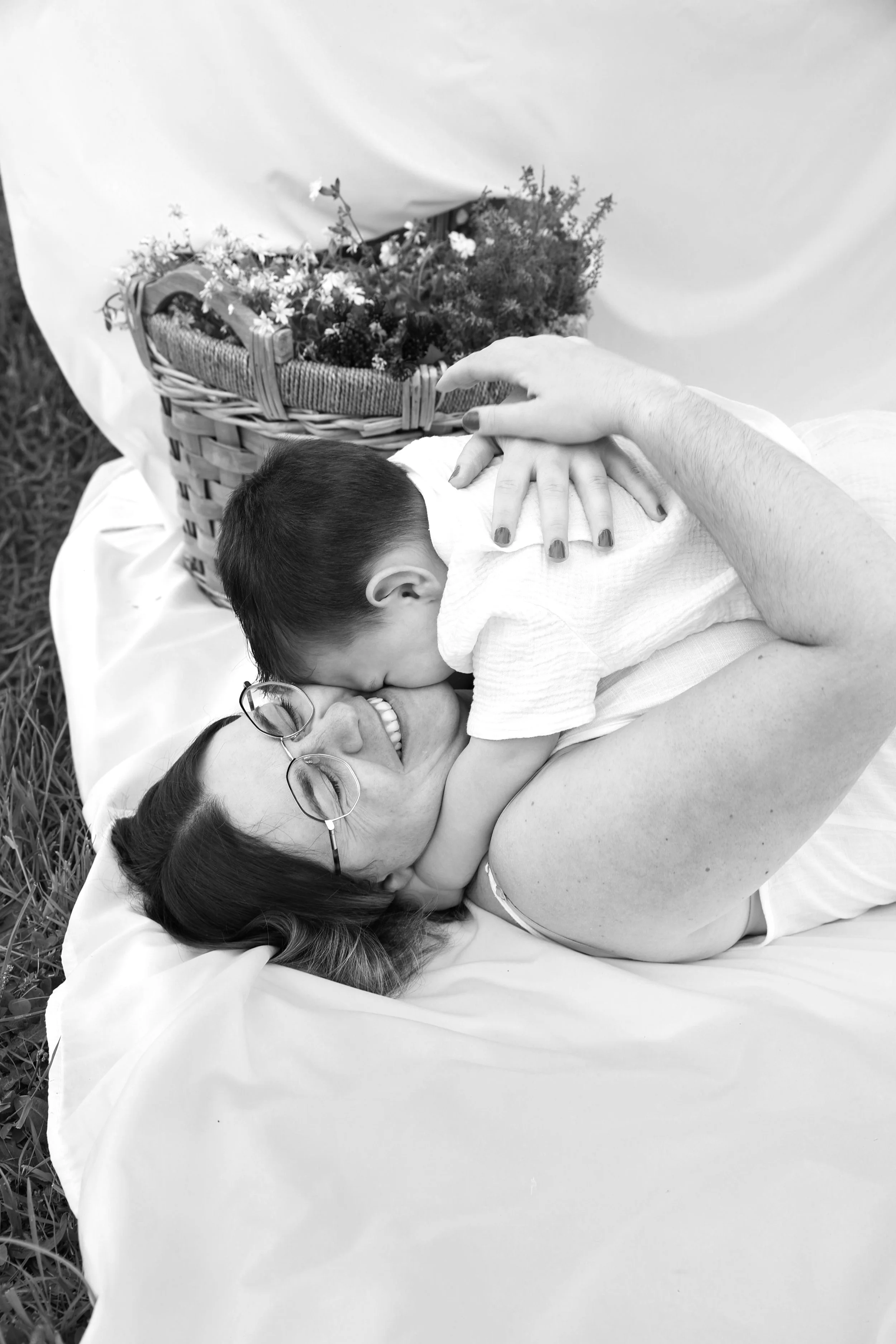 Une femme allongée sur une toile blanche à l'extérieur, entourée d'herbe, partage un moment affectueux avec un enfant qui la serre dans ses bras, avec un panier de fleurs derrière eux.