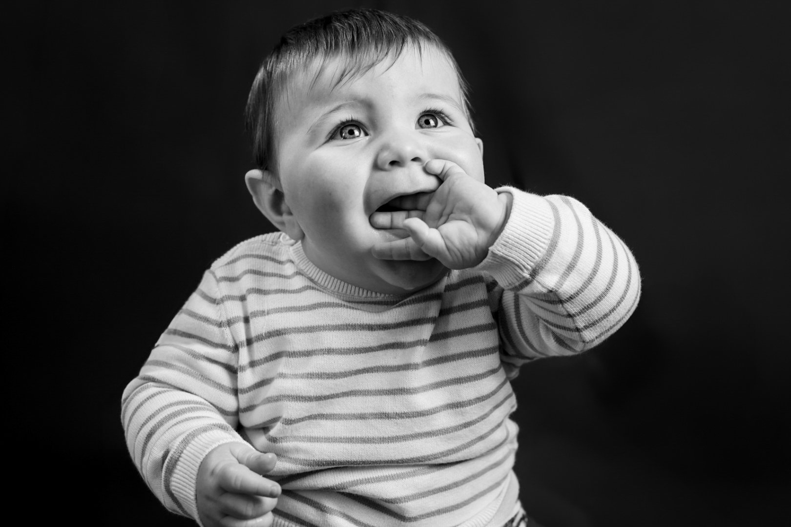 Un bébé en noir et blanc qui porte une chemise à rayures et qui se touche le visage avec la main, regardant vers le haut.