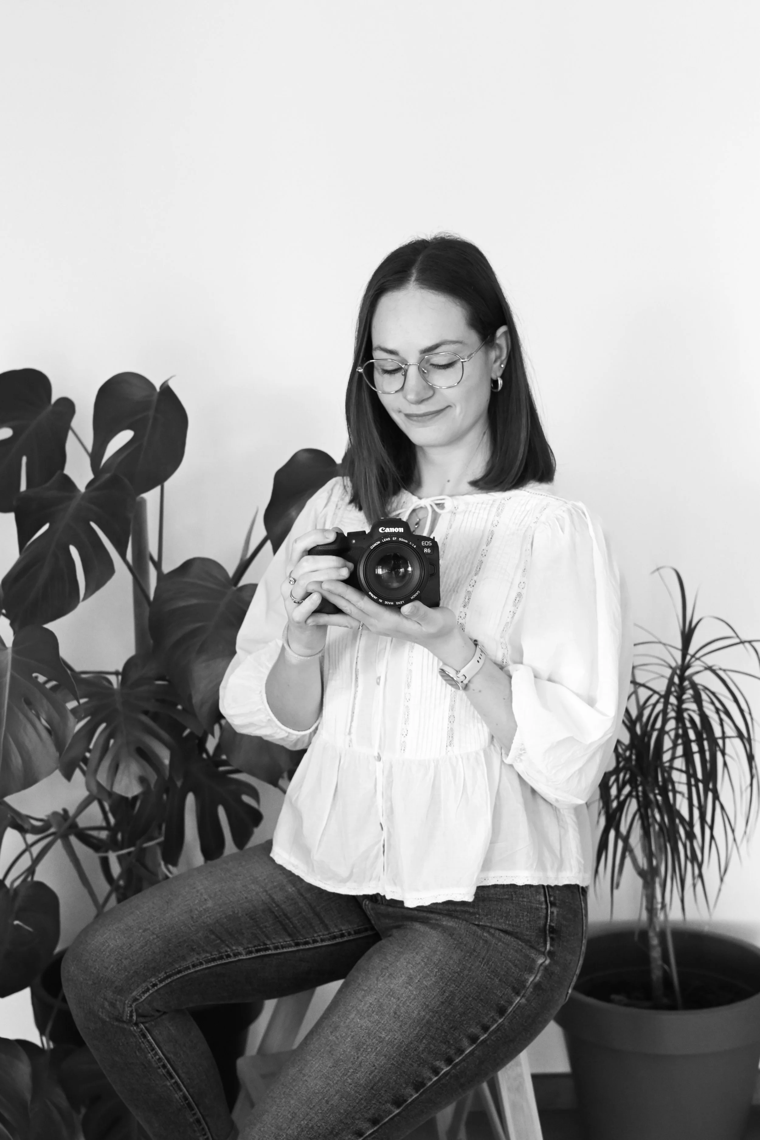 Une femme souriante, portant des lunettes, tient un appareil photo et regarde vers le bas, assise devant un fond simple avec des plantes en arrière-plan.
