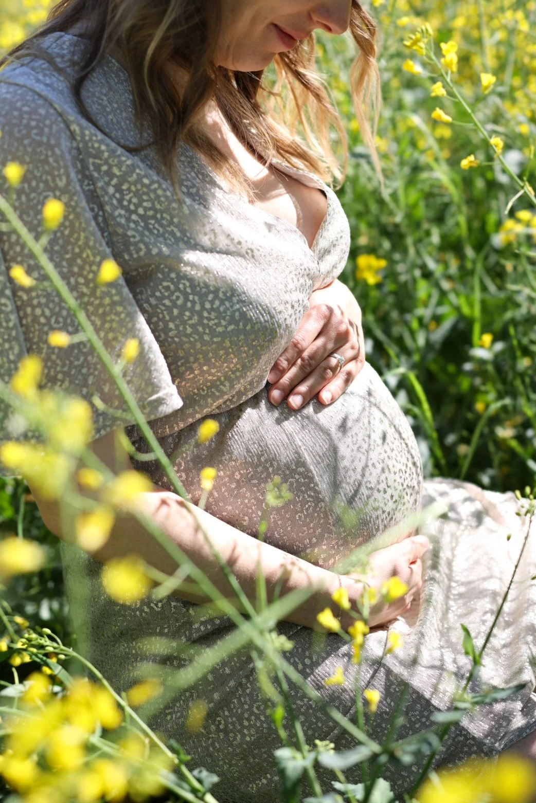 Femme enceinte assise dans un champ de fleurs jaunes, tenant son ventre avec ses mains, portant une robe claire. La scène est en plein soleil et la femme est peu maquillée.