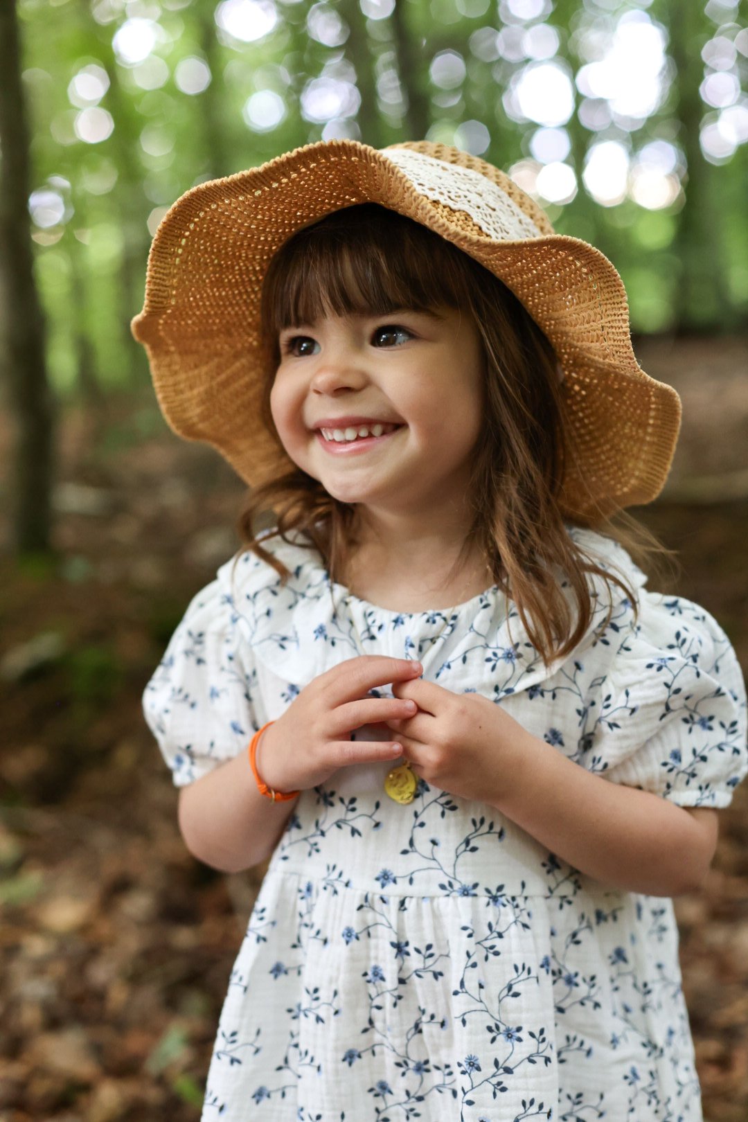 Une jeune fille souriante portant un chapeau de paille et une robe blanche avec des motifs floraux, dans une forêt verte.