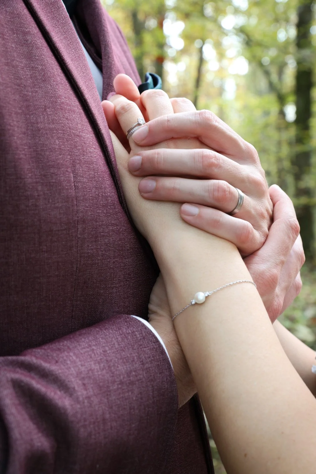 Deux personnes tenant leurs mains l'une contre l'autre devant un fond de forêt, avec des alliances et une bracelet en perle.