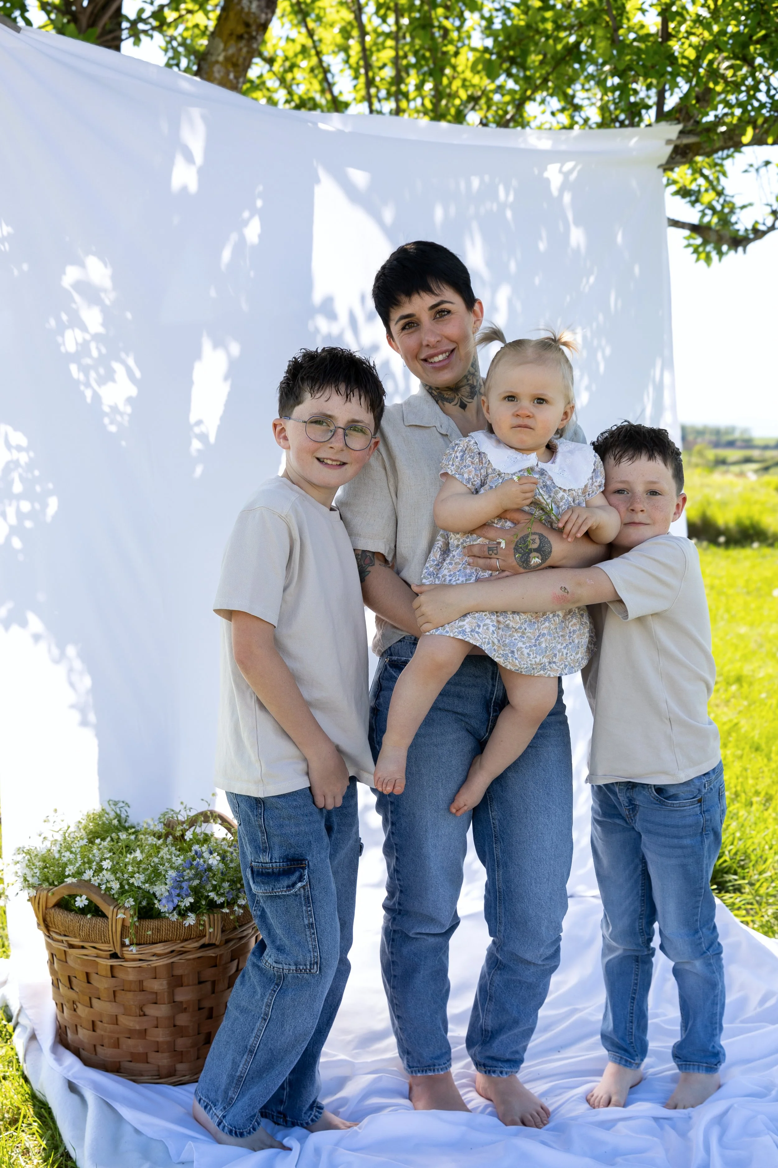 Famille posant devant un fond blanc sous un arbre, avec un panier de fleurs à ses pieds, dans un cadre extérieur verdoyant.