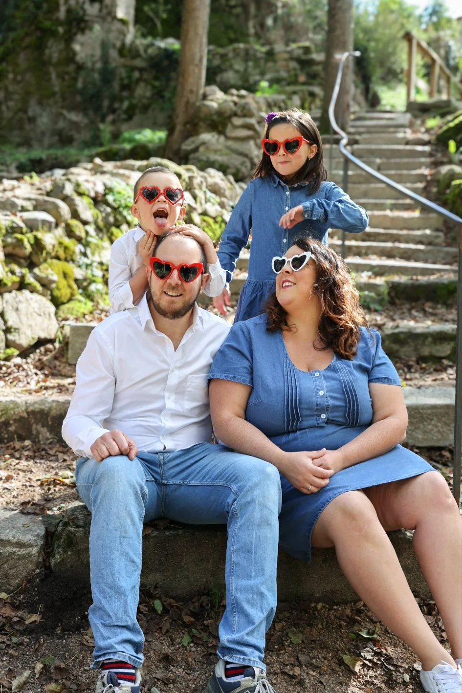 Une famille posant lors d'une séance photo en plein air, portant des lunettes de soleil en forme de cœur, avec un environnement naturel de forêt et des escaliers en pierre en arrière-plan.