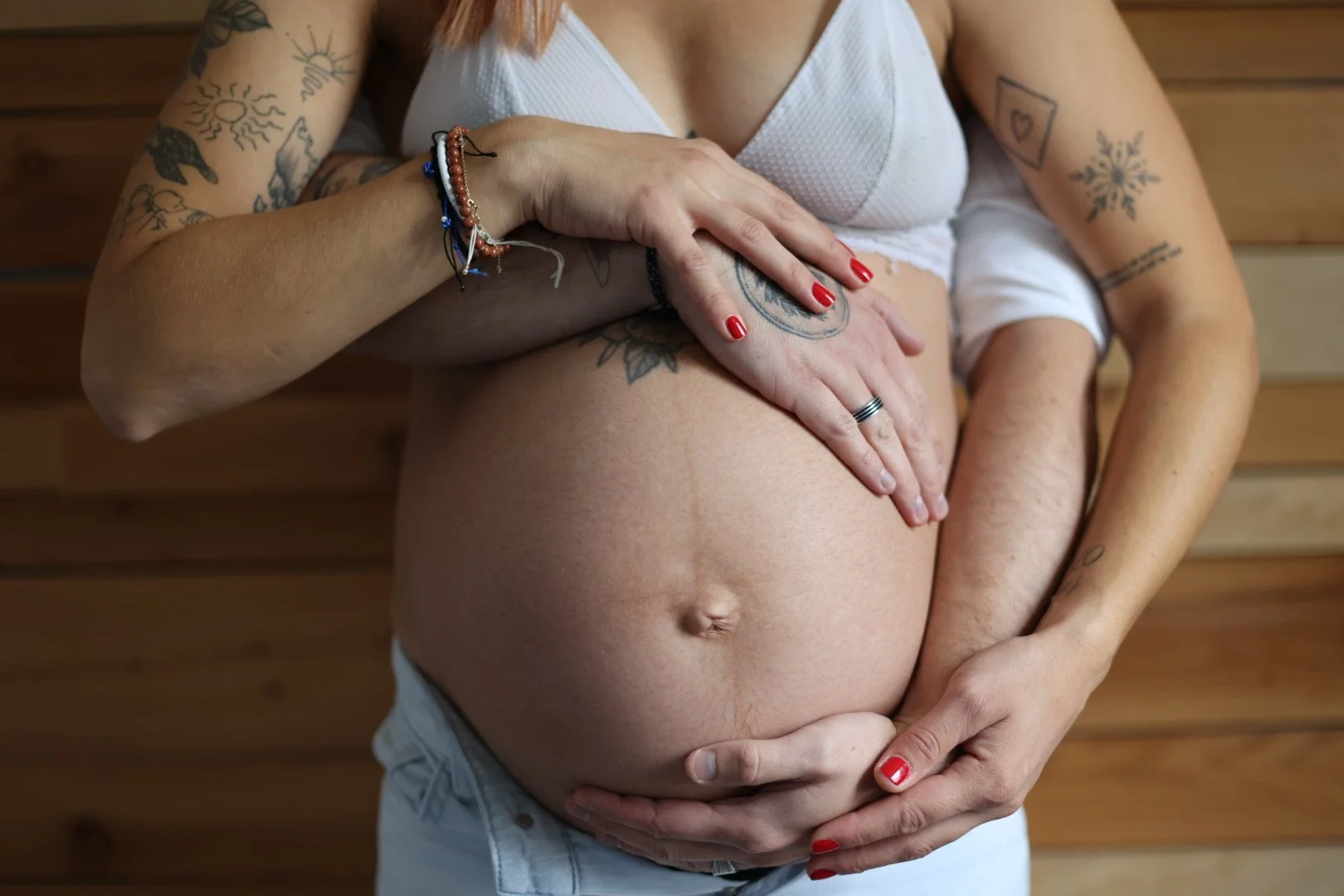 Une femme enceinte avec plusieurs tatouages, tenant son ventre arrondi avec ses mains. Elle porte des ongles rouges et des bracelets. Le fond est en bois.
