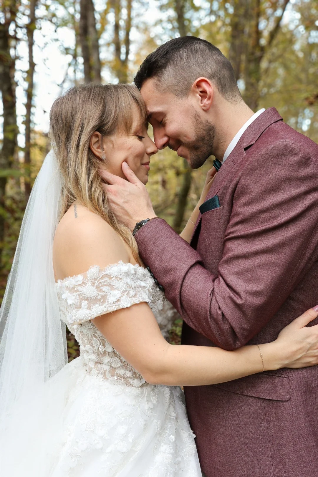 Un couple de mariés, un homme et une femme, se tiennent face à face en forêt, le front et le nez pressés l'un contre l'autre, tenant le visage de l'autre avec tendresse, lors d'une séance photo de mariage.