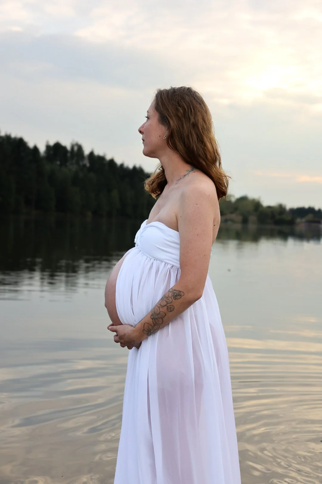Femme enceinte en robe blanche, debout près d'un lac, regarder à gauche, ambiance tranquille au coucher du soleil.