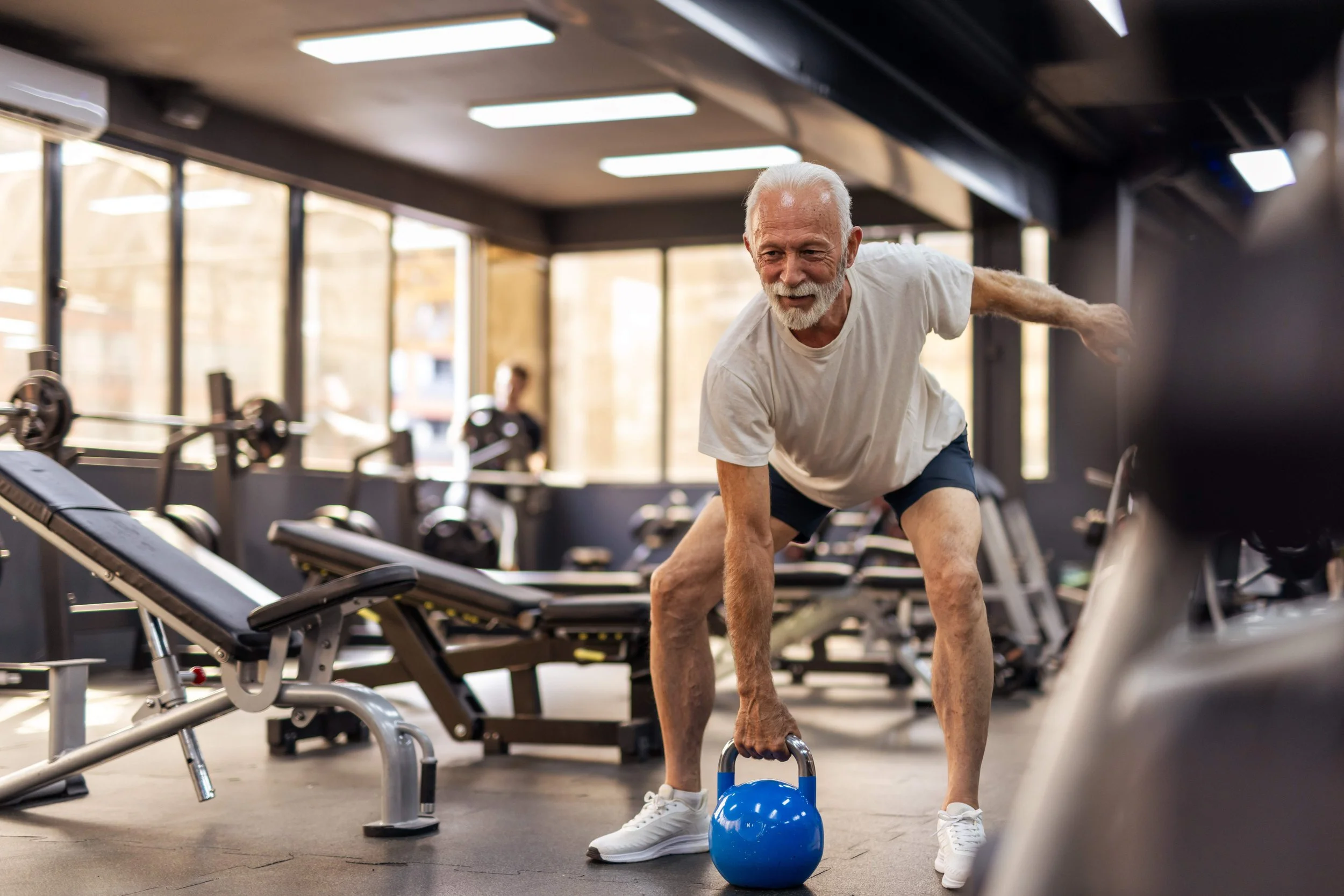An elderly man with white hair and a white beard working out in a gym. He is performing a kettlebell exercise using a blue kettlebell, wearing a light gray t-shirt, dark shorts, and white sneakers. The gym has large windows, various exercise equipment, and a person in the background.