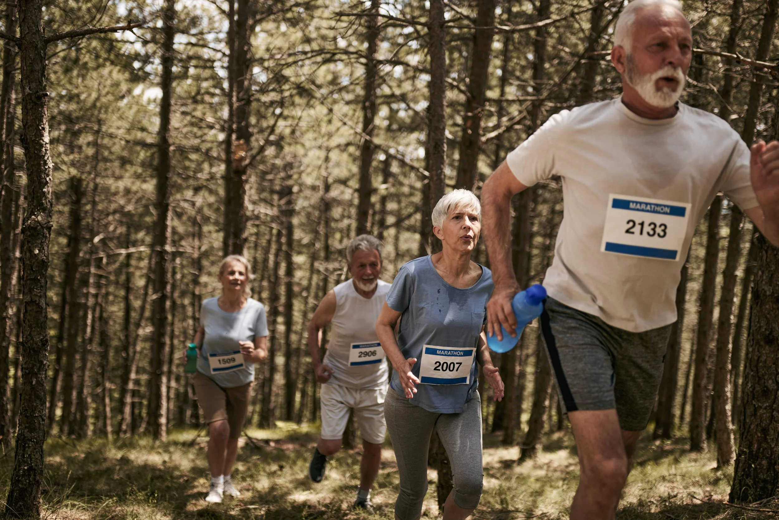 A group of older adults running or jogging in a forest during a marathon event, wearing race bibs and holding water bottles.