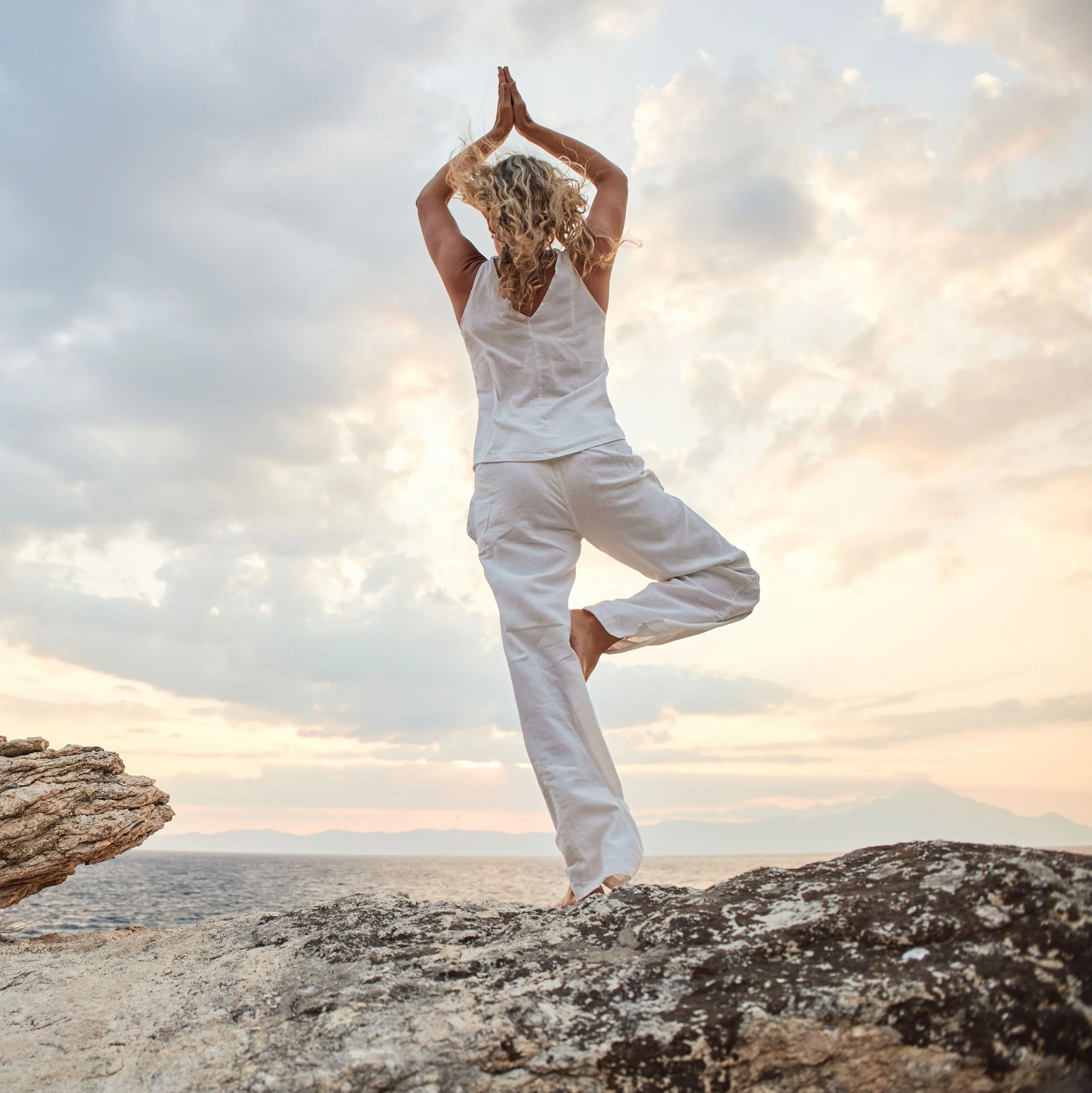 A woman practicing yoga on a rocky ledge by the ocean at sunset, balancing on one leg with arms raised and palms pressed together overhead.