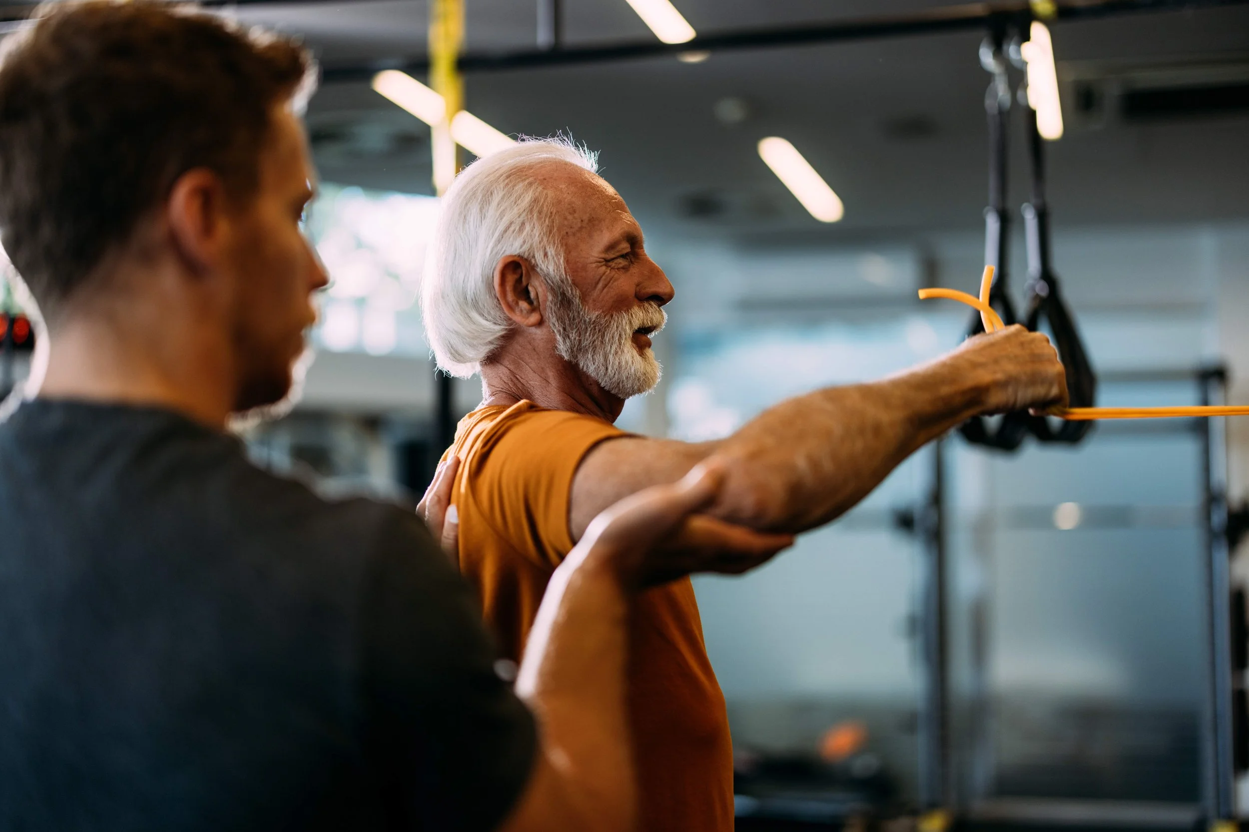 The photo shows two men in a gym. One is an older man with a white beard and hair, smiling and engaging in a workout. The other man, closer to the camera, has darker hair and is slightly out of focus. The older man is holding a resistance band or similar exercise equipment.