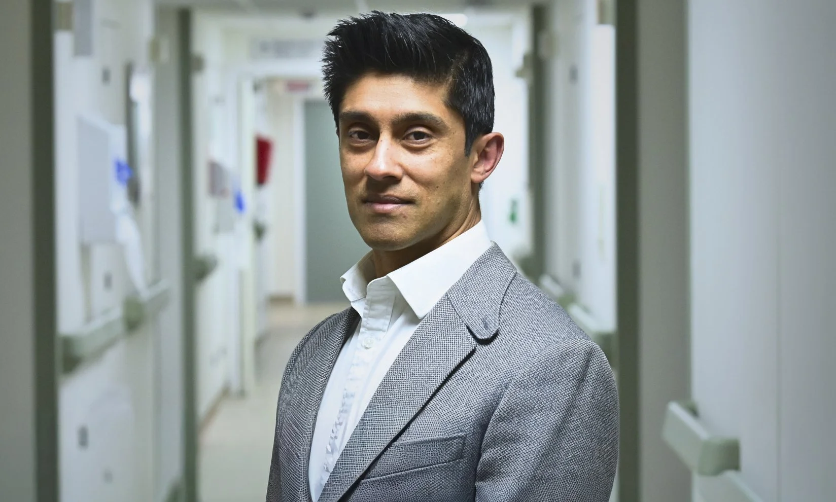 Portrait of a man in a gray blazer and white shirt standing in a hallway lined with lockers.