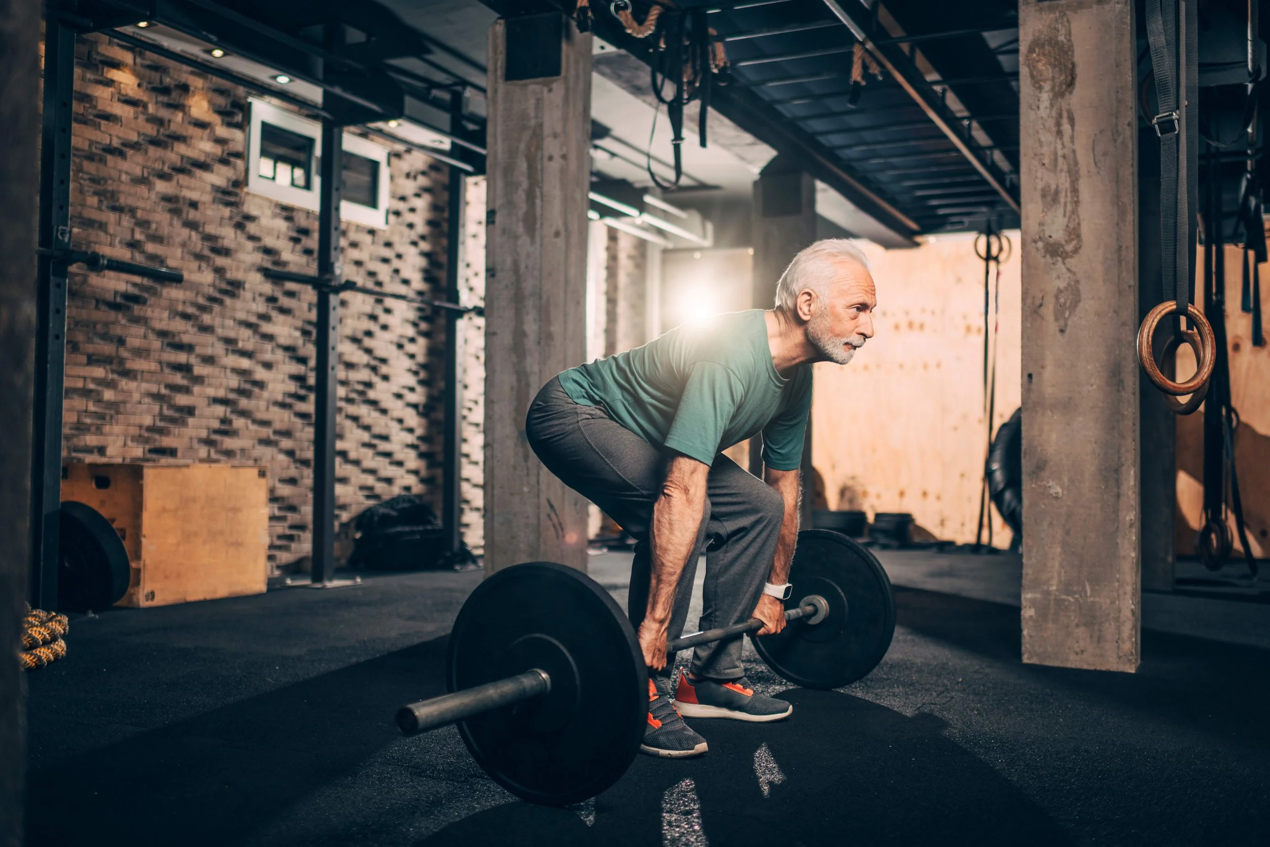 An elderly man with gray hair and a beard lifting a barbell in a gym with exposed brick walls and workout equipment.