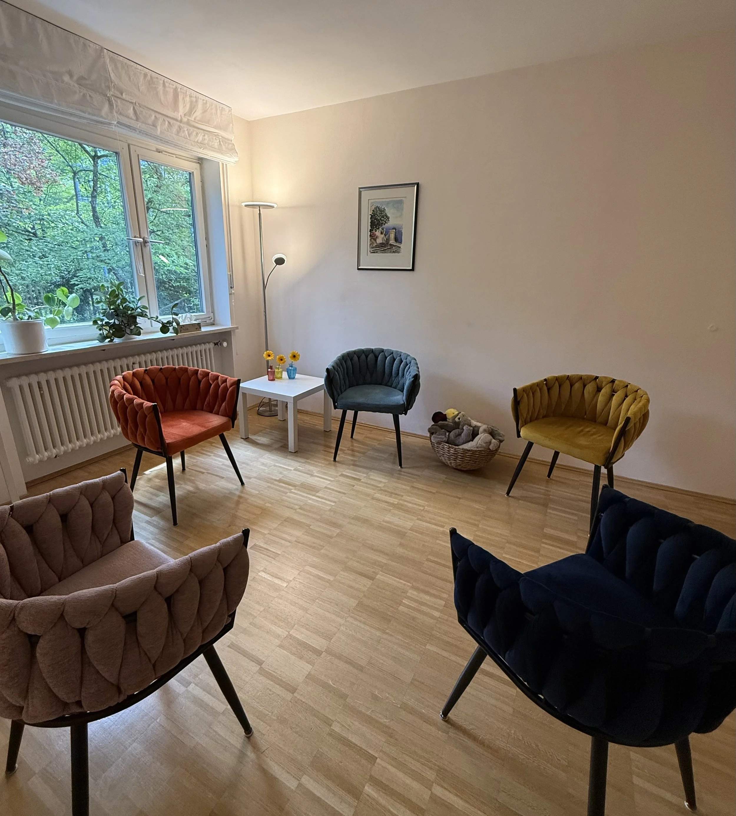 Innenraum mit mehreren bunten Sesseln, an einem weißen Tisch mit kleinen Vasen und Sonnenblumen, einer Fensterfront mit grünen Bäumen im Hintergrund, einer Stehlampe und einem Wandbild.