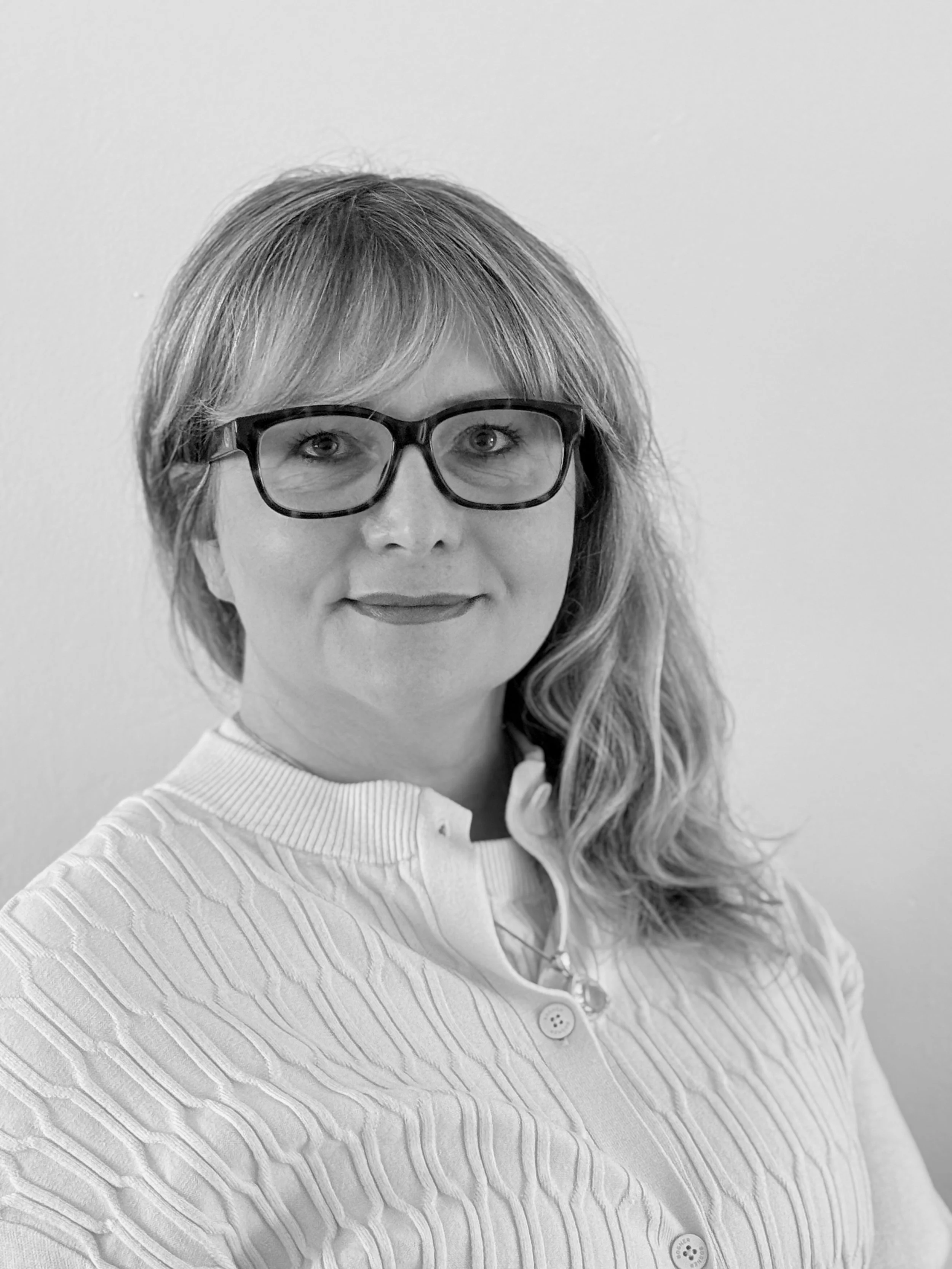 Schwarz-weiß-Bild einer Frau mit Brille, schulterlangem Haar, lächelnd, in einem weißen, gerippten Kleid, vor neutralem Hintergrund.