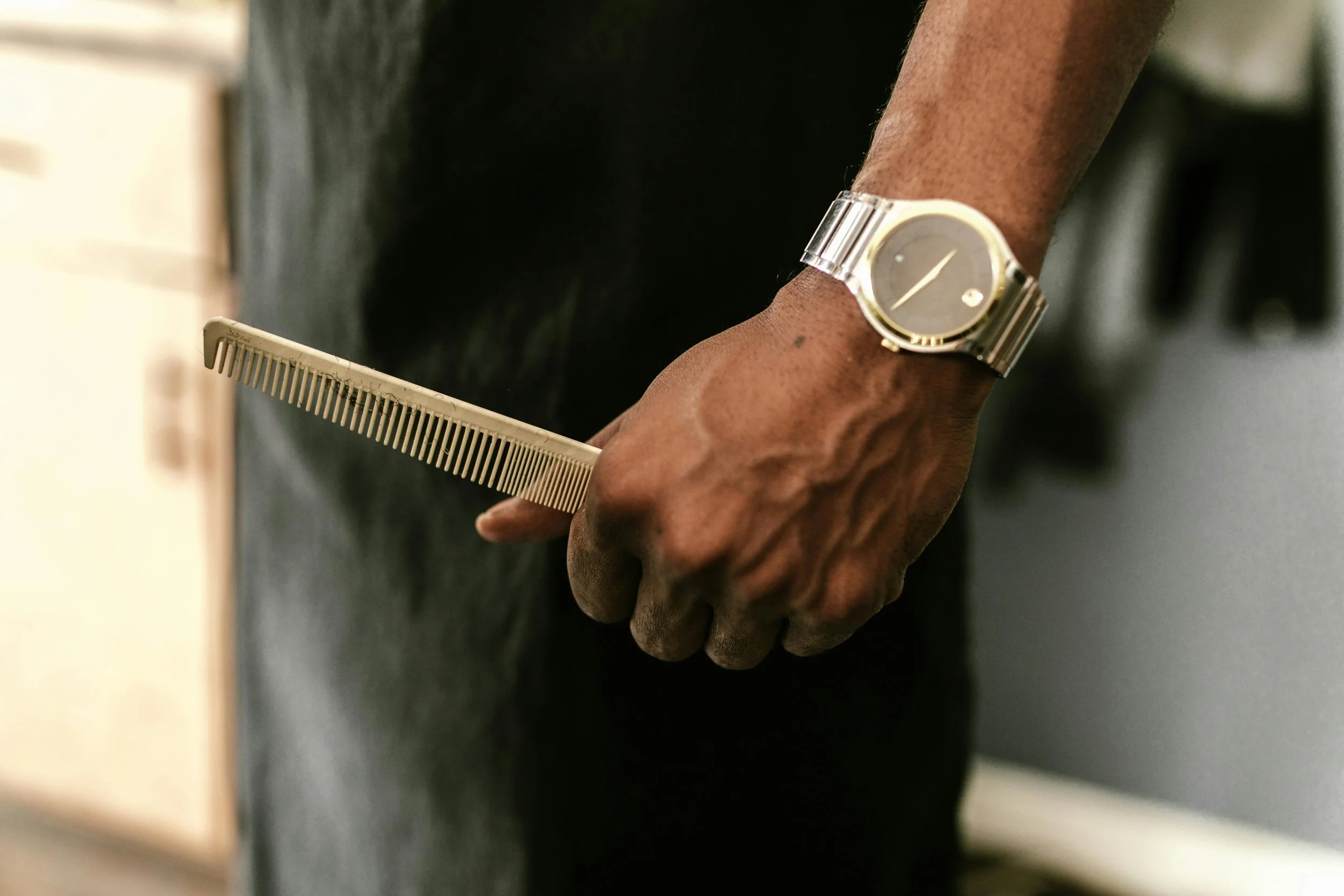 Close-up of a person's hand holding a silver comb, wearing a silver wristwatch, with a dark background.
