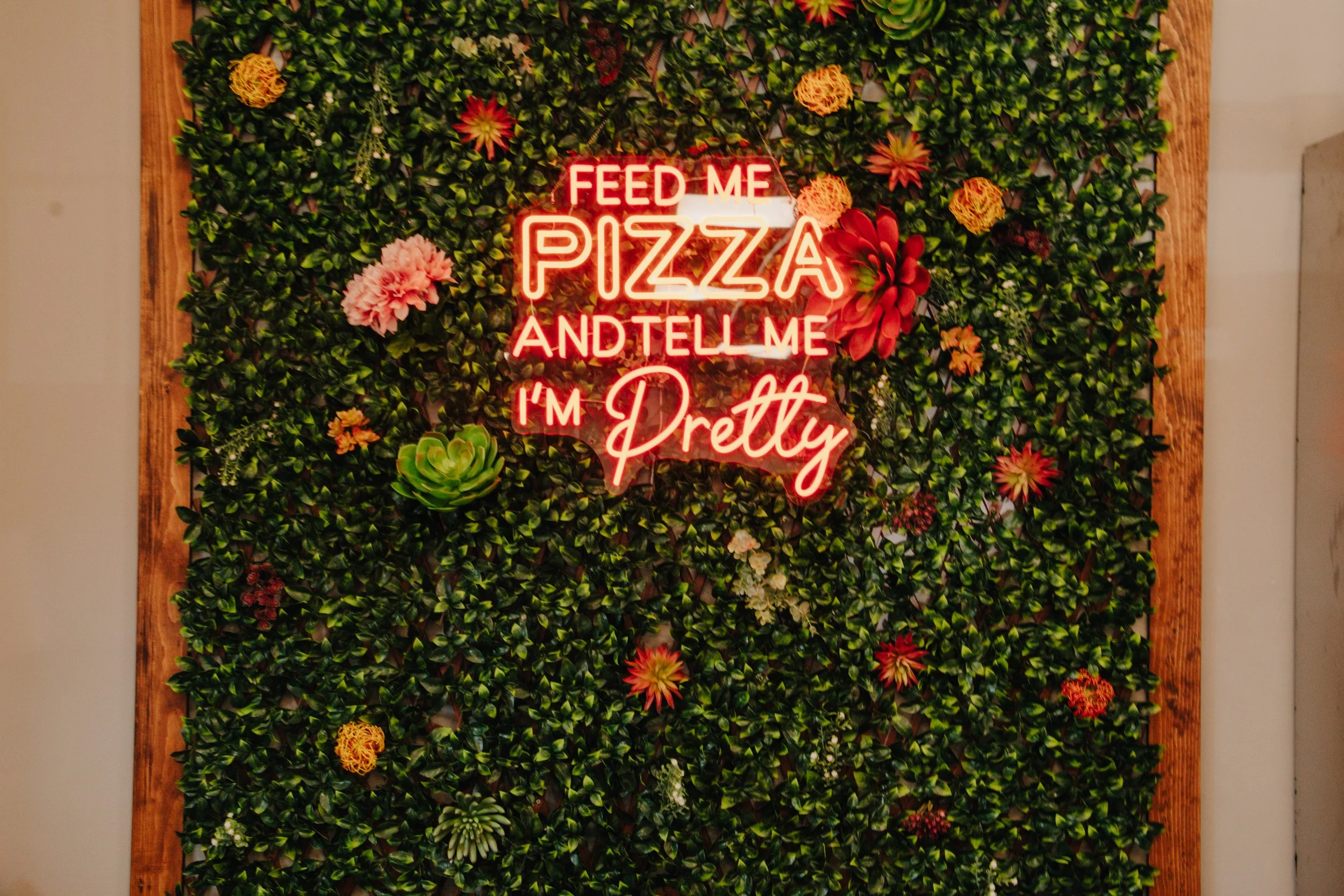 A decorative wall with green foliage, colorful flowers, and a neon sign that says, 'FEED ME PIZZA AND TELL ME I'M PRETTY'.