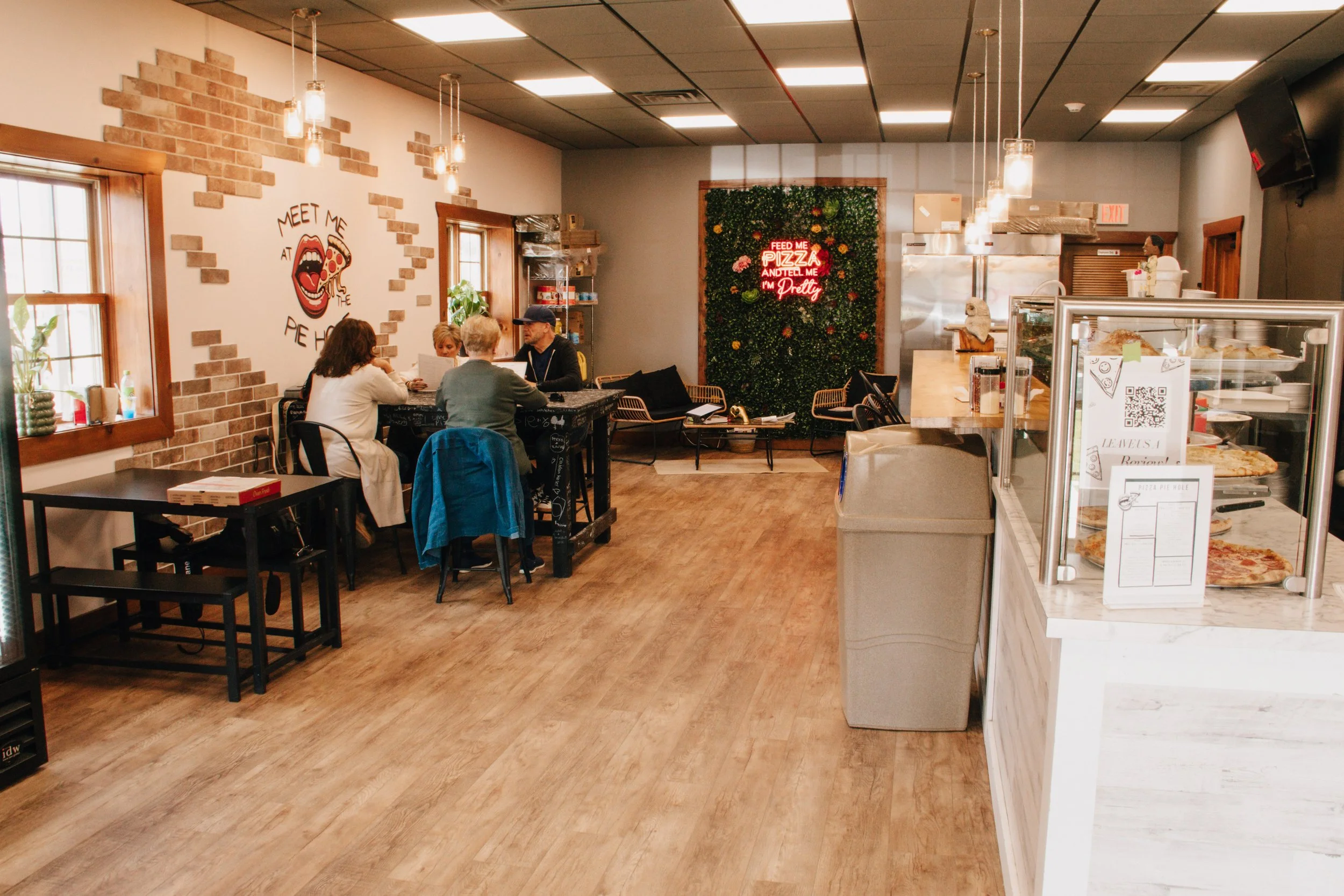 Inside a pizza restaurant with a few customers sitting at a table and a display case with pizzas on the right. Decor includes brick wall accents, a green wall with flowers, and neon signs.