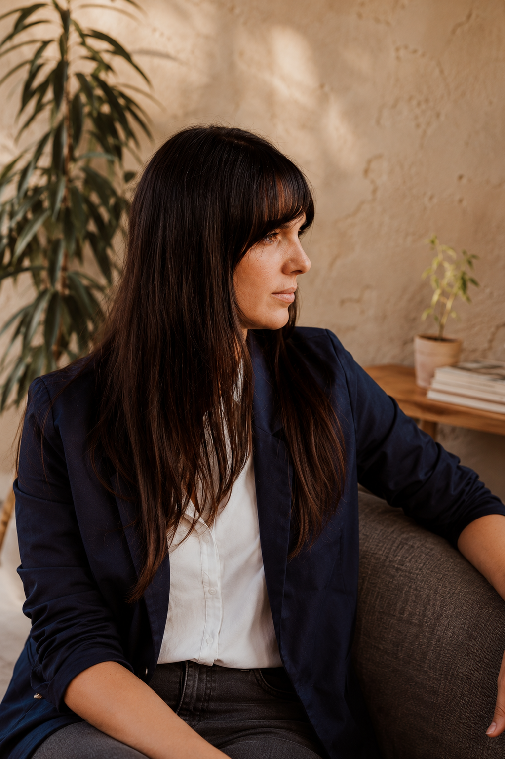 A woman with long dark hair and bangs, wearing a white blouse and navy blazer, sitting indoors with plants and a wooden table in the background.