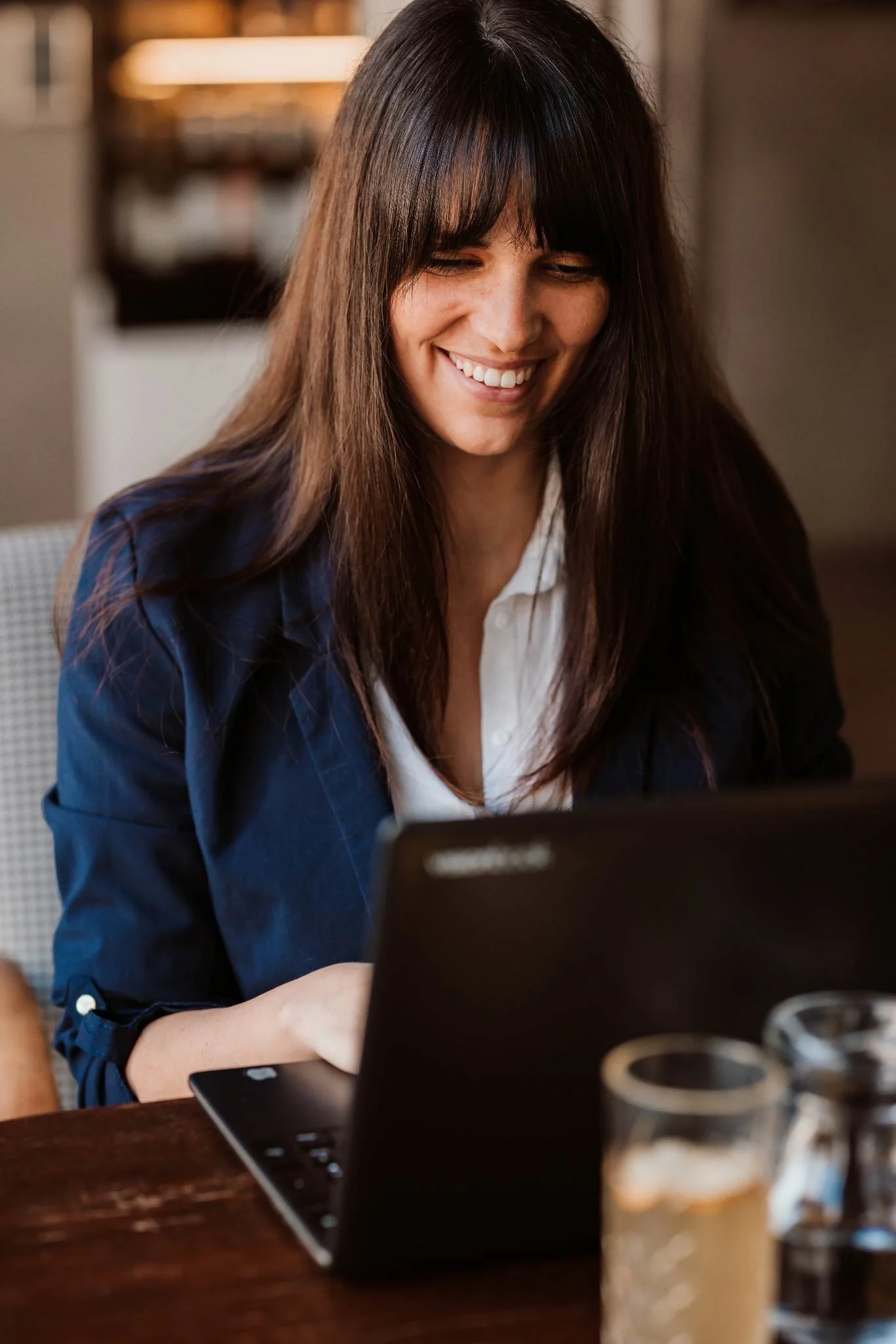 A woman with long dark hair and bangs is smiling while looking at a laptop in front of her. She is sitting at a wooden table with glasses of beer in the foreground. The background appears to be indoors, possibly in a cafe or office with warm lighting.