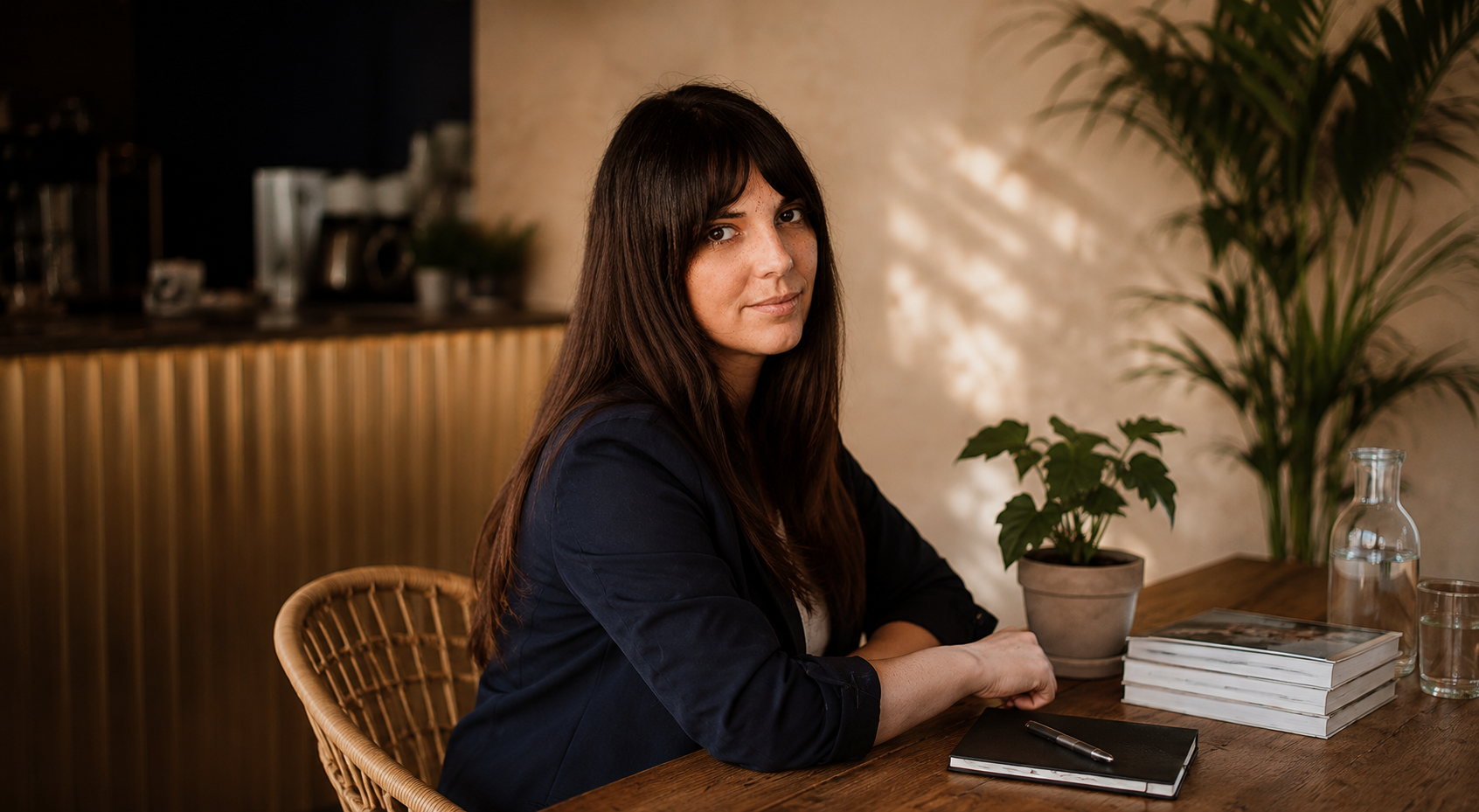 A woman with long dark hair and freckles sitting at a wooden table with a stack of books, a potted plant, a notebook, a pen, and glasses of water in a warmly lit cafe or restaurant.