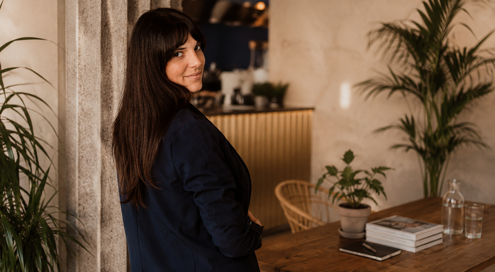 A woman with long dark hair, wearing a black blazer, standing in a cozy, stylish room with plants and a wooden table with books and glasses.