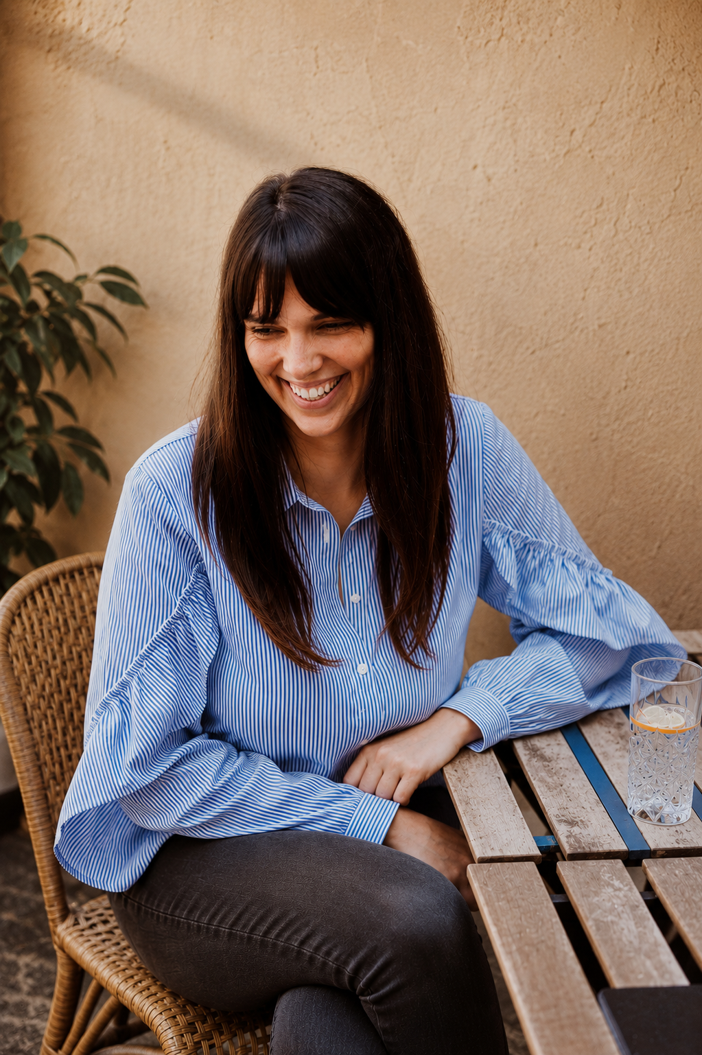 A woman with long dark hair wearing a blue and white striped shirt, sitting at a wooden table outdoors, smiling and looking down, with a glass of water nearby.