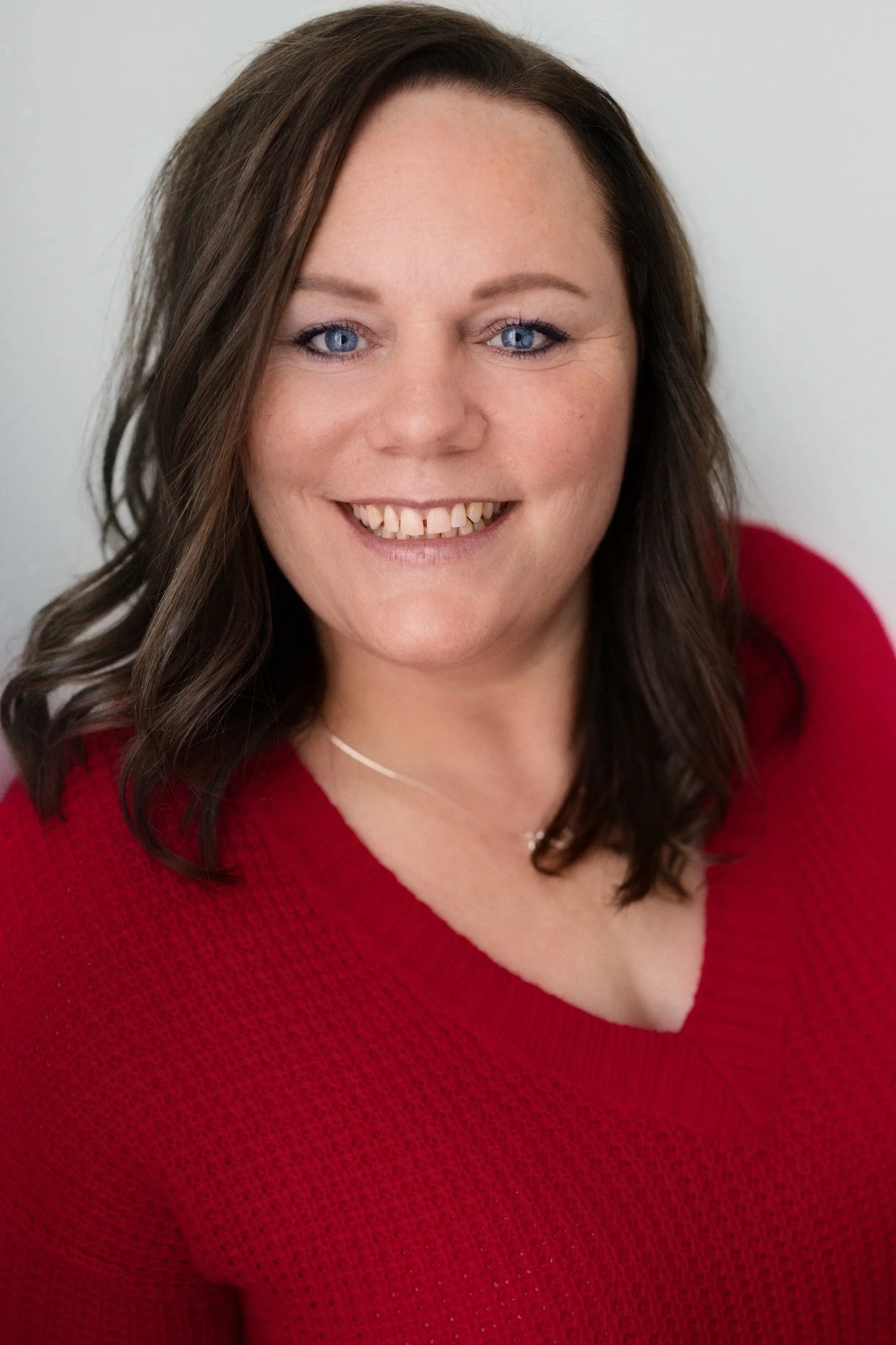A woman with brown hair, blue eyes, and a wide smile, wearing a red V-neck sweater and a thin necklace, against a plain light background.