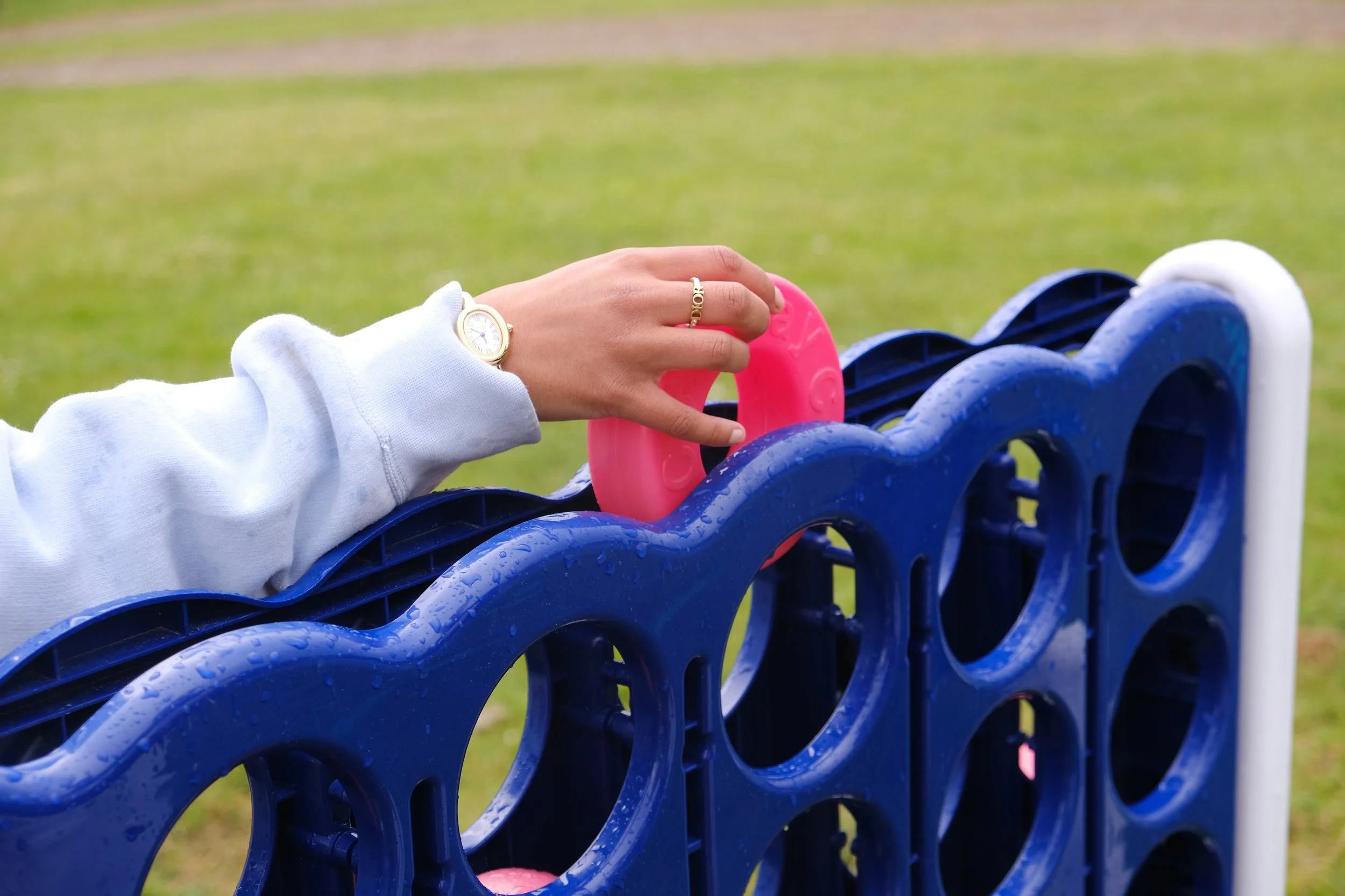A person's hand wearing a gold watch and a ring is reaching to grab a pink object on a blue circular structure outdoors on a rainy day.