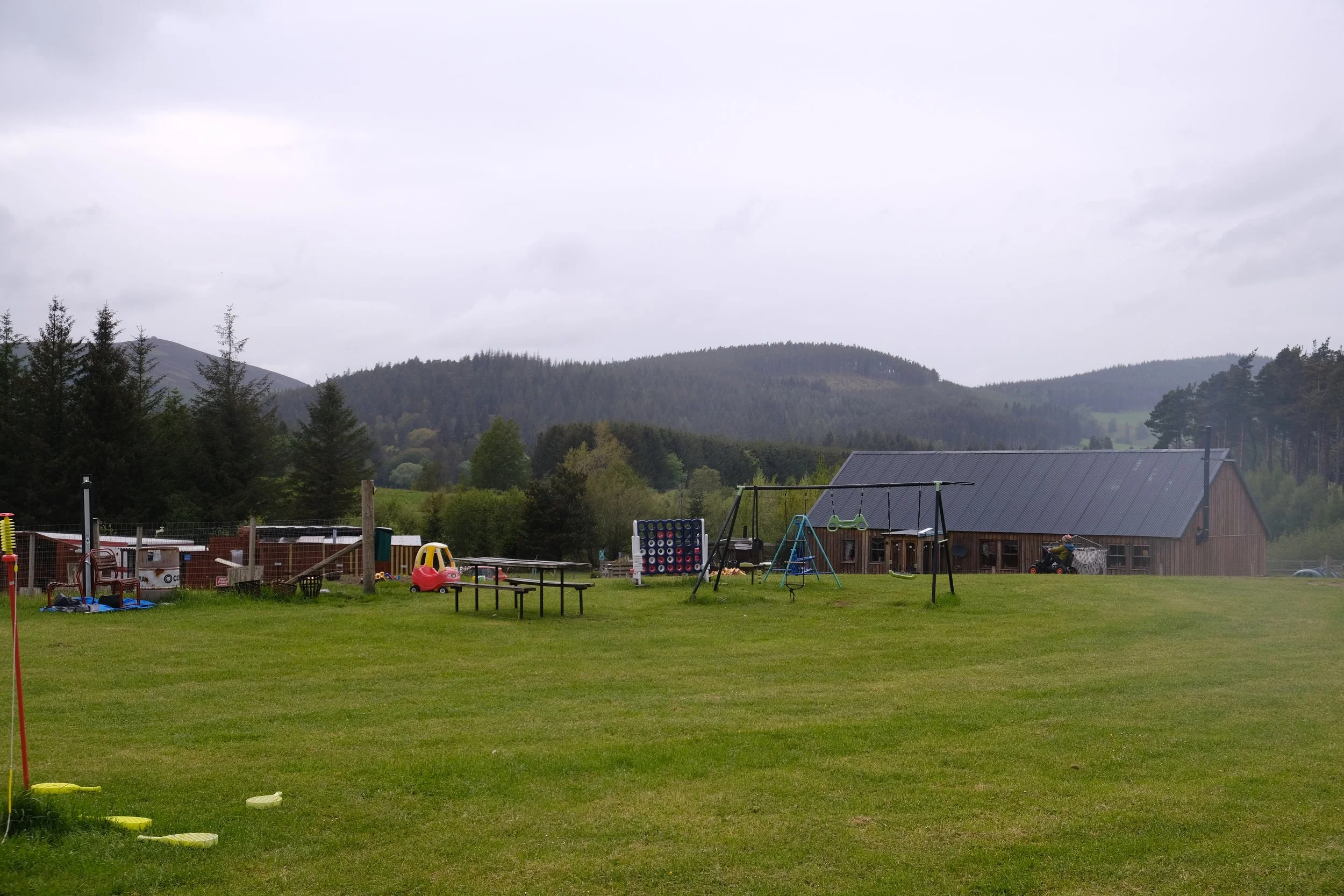 A grassy outdoor playground with toys and playsets, surrounded by a fence. In the background, there are houses, trees, and mountains under a cloudy sky.