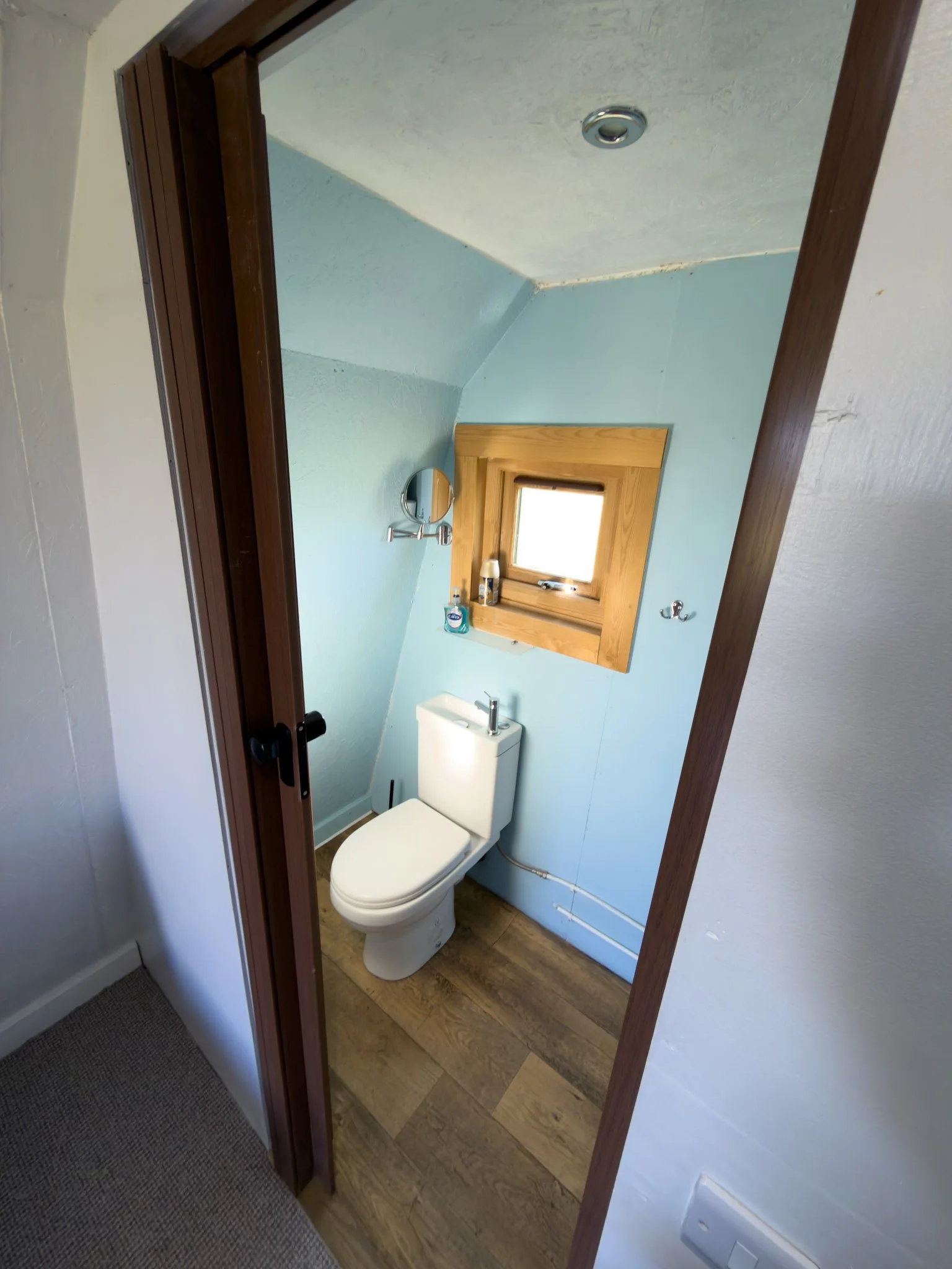 A small bathroom with a white toilet, a wooden framed window, a small mirror, and a vanity with toiletries, seen through a doorway with a wooden trim.
