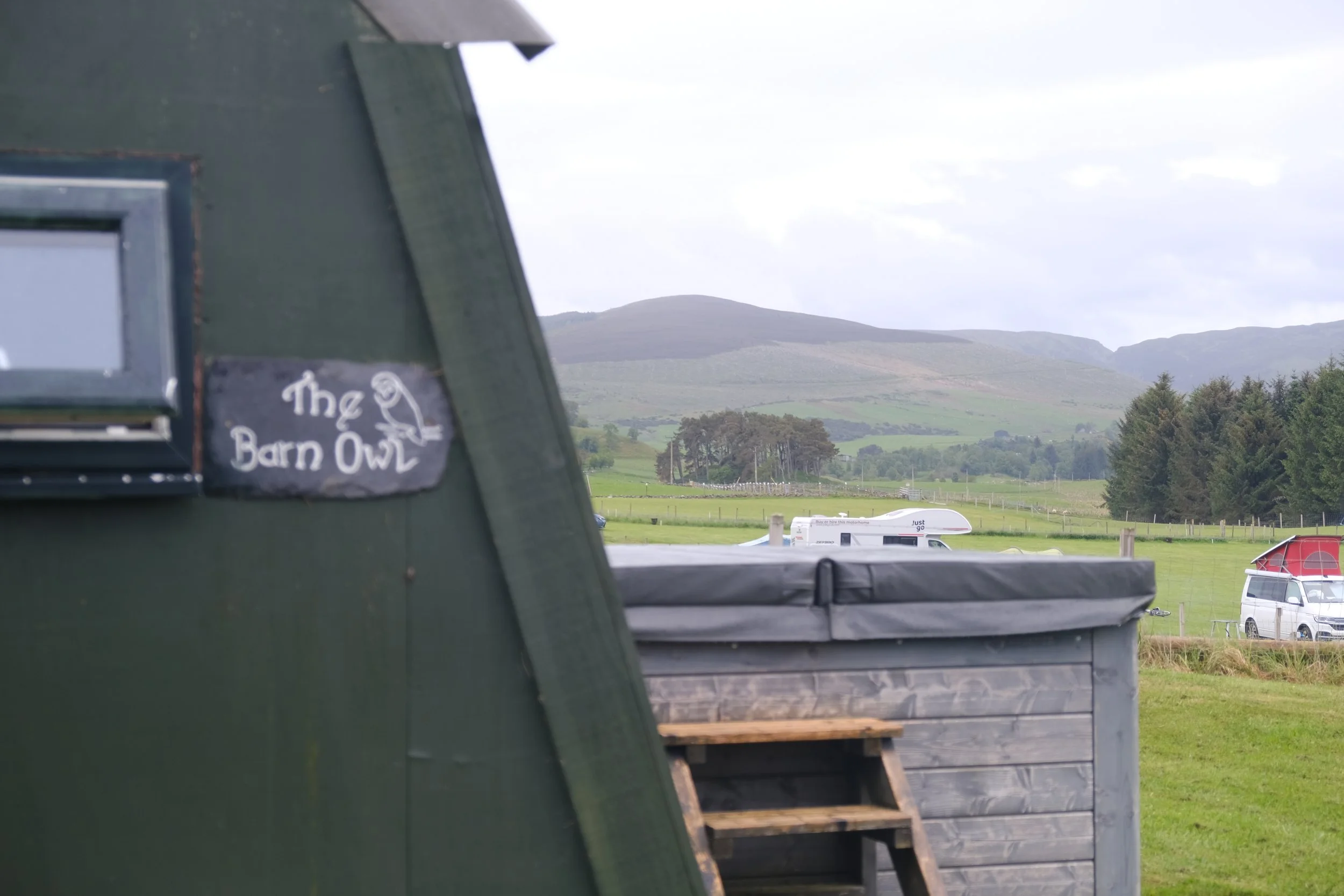 View of a rural landscape with green hills, trees, and cloudy sky, seen through a partially open window or opening with a collage of various objects, including a sign that reads 'The Barn Owl'.