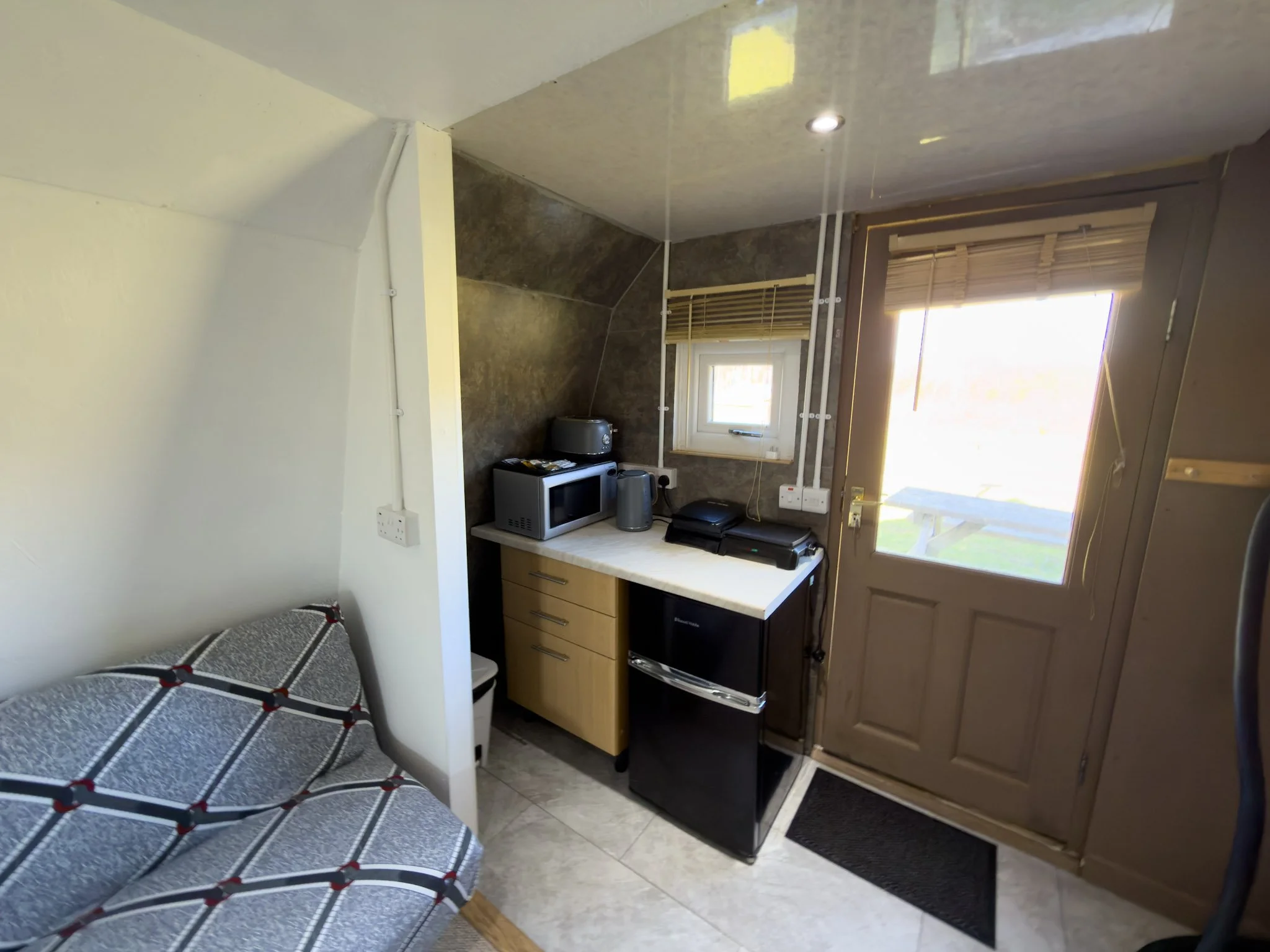 A small kitchen with a black stove, microwave, toaster, and small fridge on a beige countertop. There is a window with wooden blinds and a door with glass panels. The wall is covered with dark brown tiles.