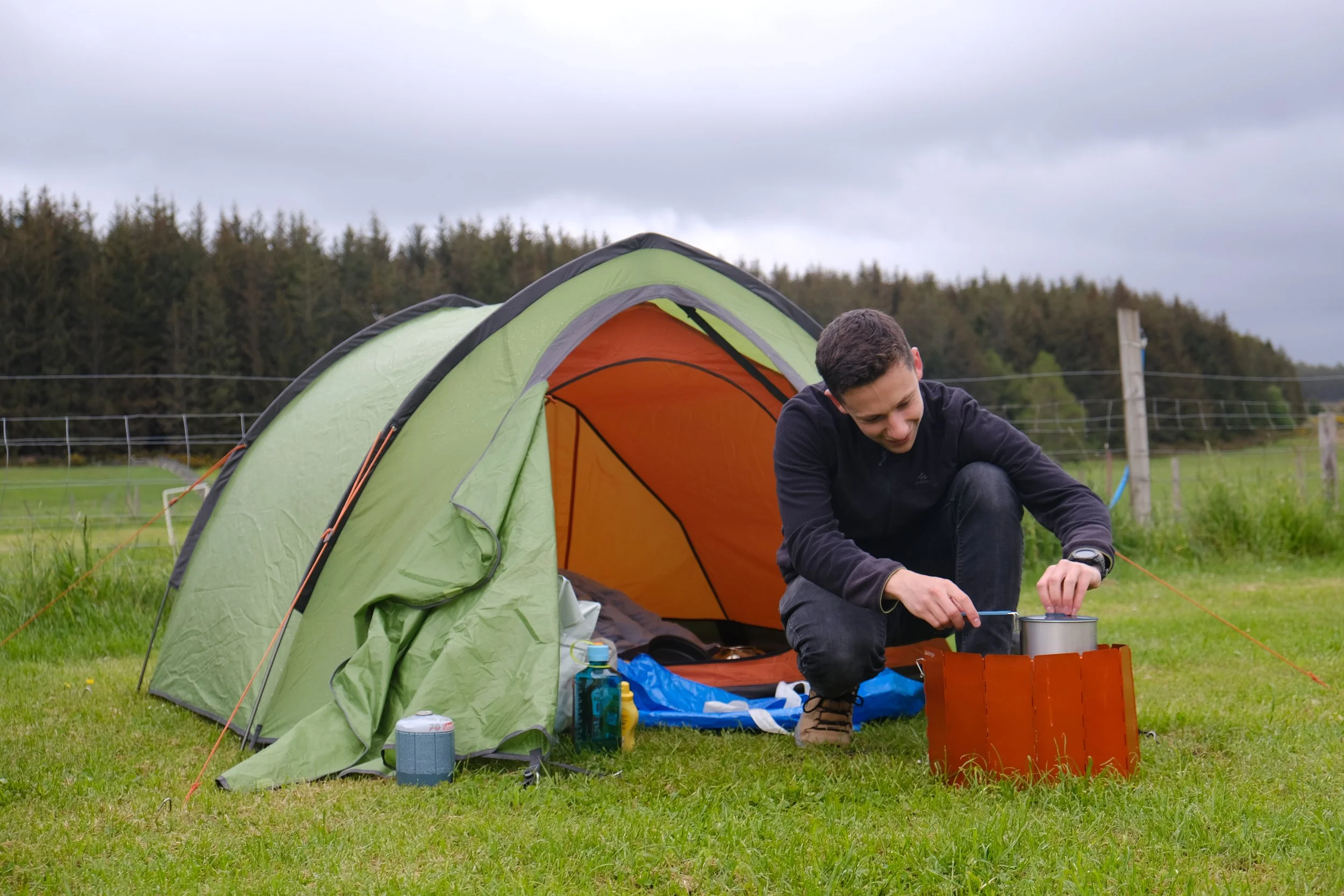 A man preparing camping gear outside a green tent in a grassy field with trees and gray cloudy sky in the background.