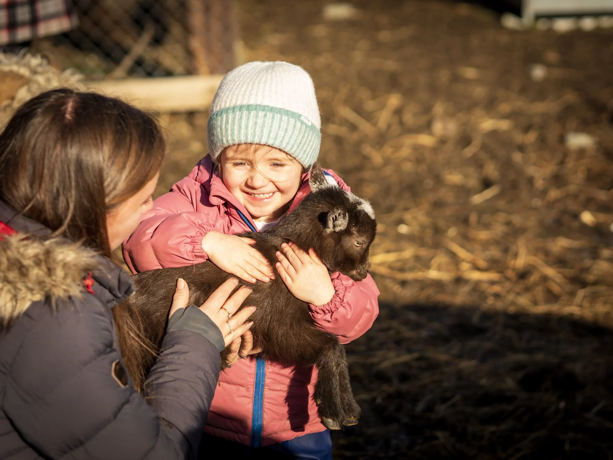 A young girl in a pink jacket and a white beanie holding a small puppy, smiling, with an adult woman in a dark jacket and a fur-lined coat nearby, outdoors in a natural setting.