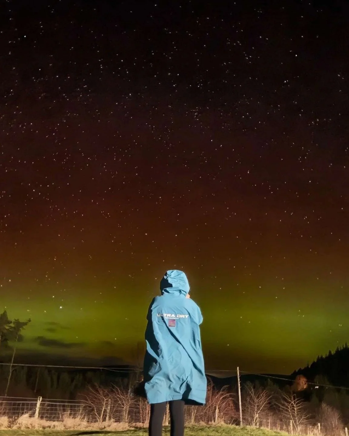 A person in a blue jacket standing outdoors at night, looking up at a starry sky with green aurora in the background.