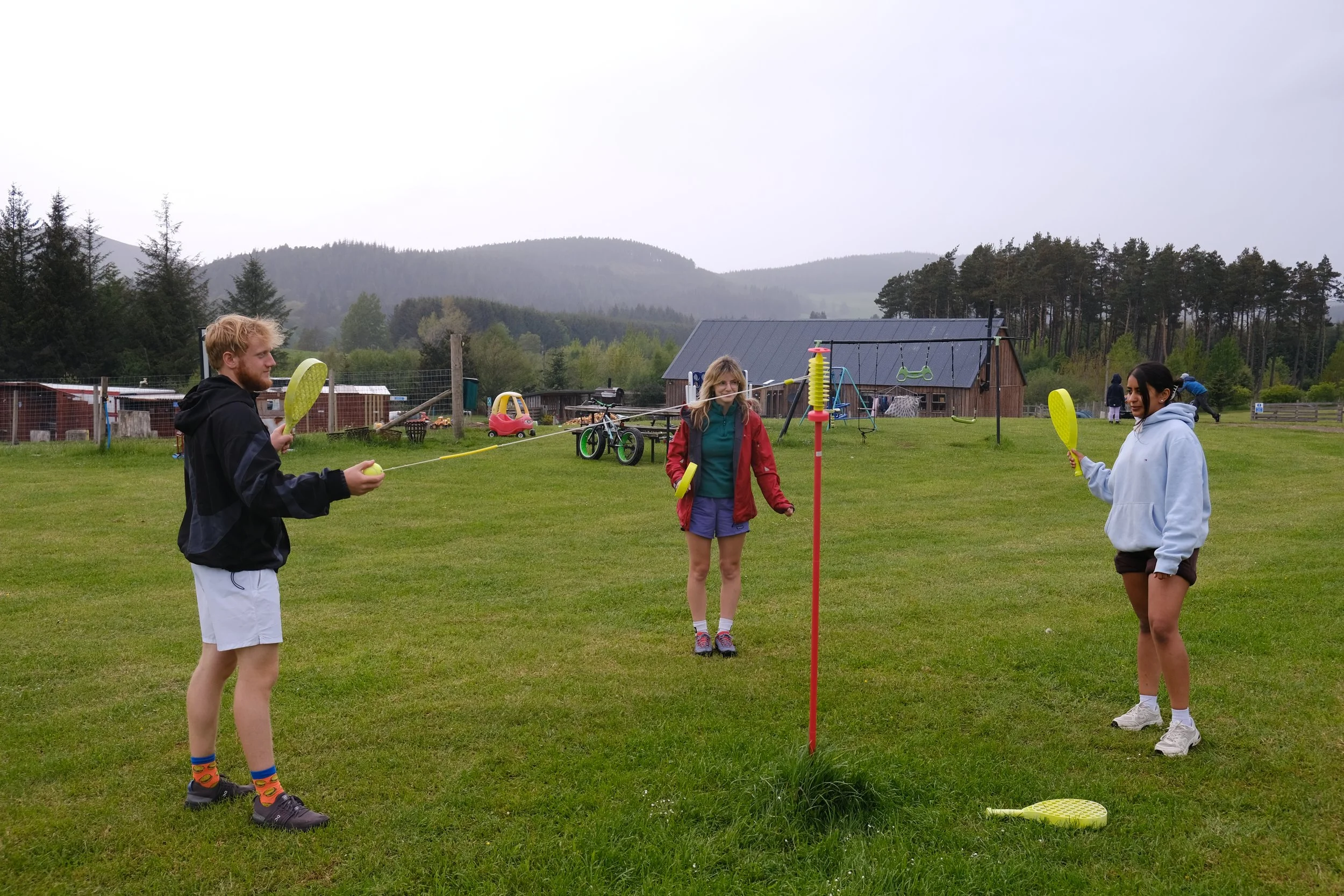 Three people playing a game with a yellow ball and rackets on a grassy field, with a swing set, a barn, and mountains in the background.