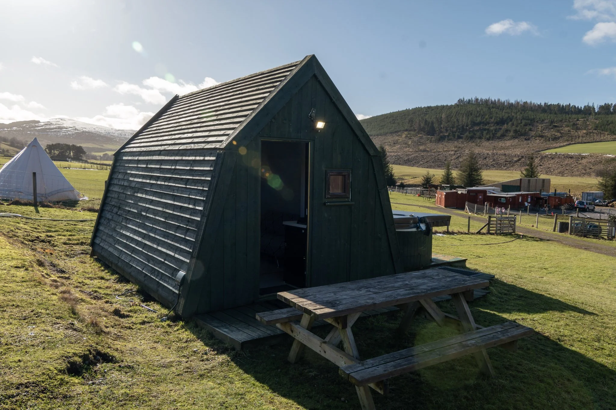 Small dark wooden building with a steep roof and small window, set on a raised wooden platform in a grassy field, with similar structures and rolling hills in the background under a blue sky with some clouds.