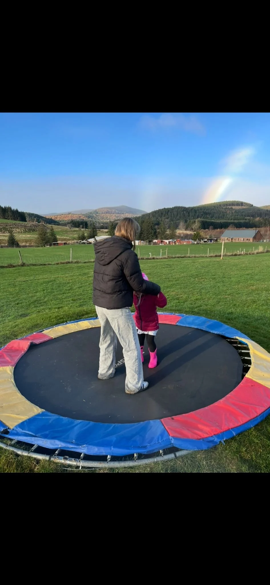 A woman and a young girl stand on a trampoline outdoors in a large green field with rolling hills and a rainbow in the sky.