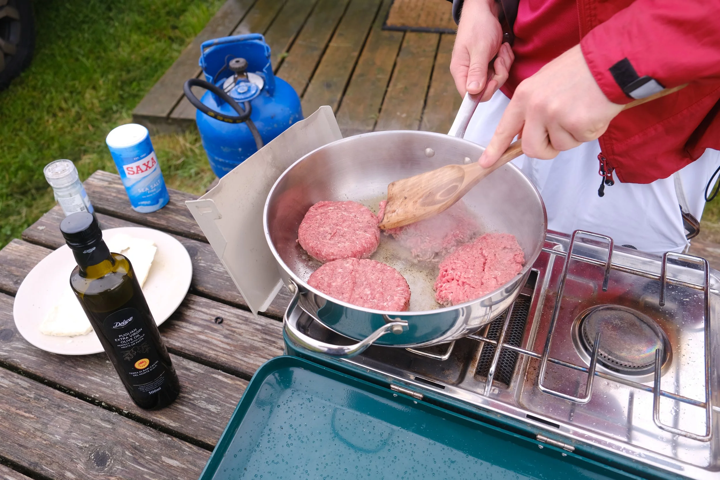 Person cooking hamburger patties on a portable stove outdoors, with a propane tank, salt container, and olive oil bottle nearby on a wooden table.