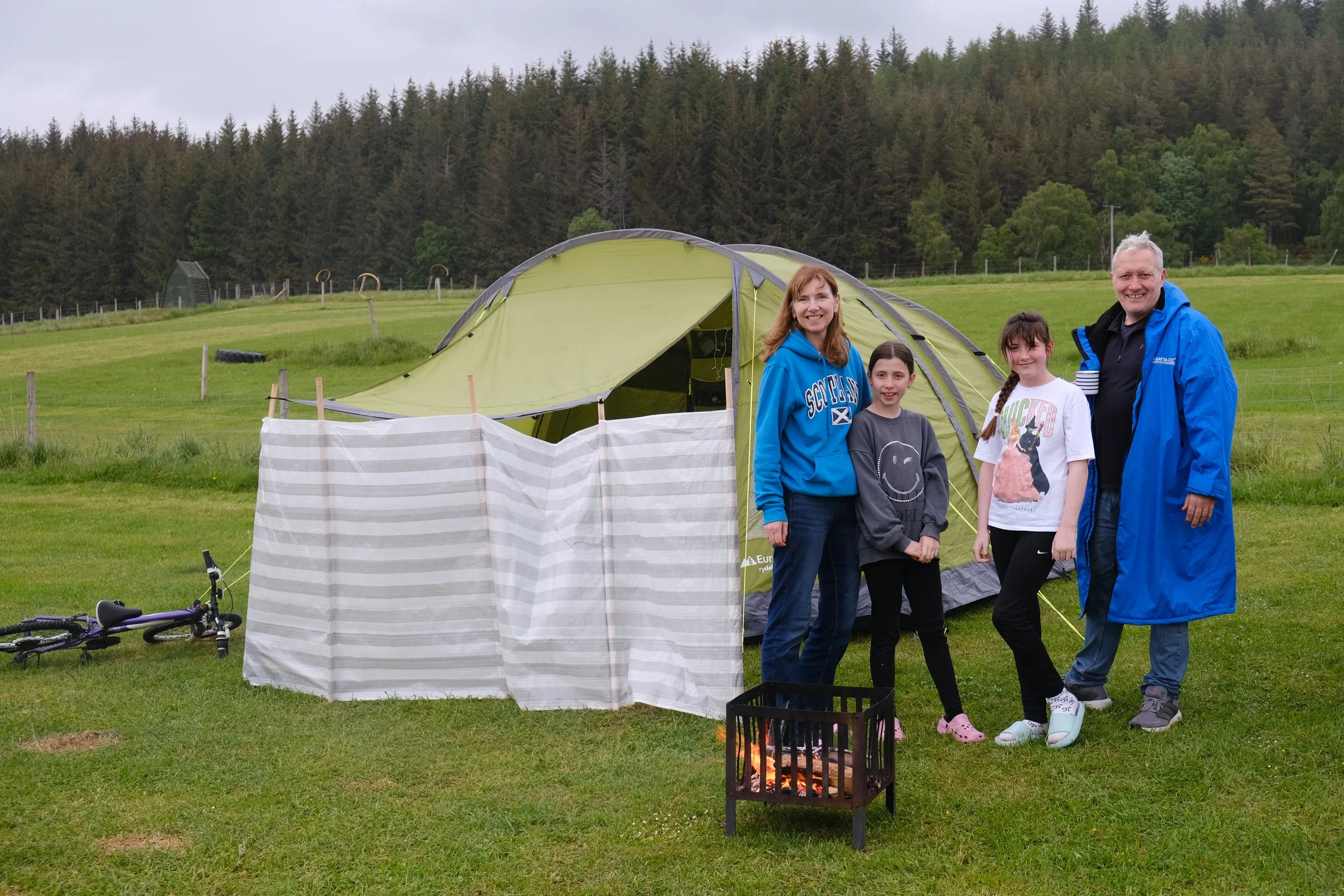 A group of four people standing outdoors near a green camping tent with a forested hillside in the background. They are smiling and dressed casually, with some wearing jackets. A small campfire is burning in front of them, and a bicycle and a clothes drying rack are visible nearby.