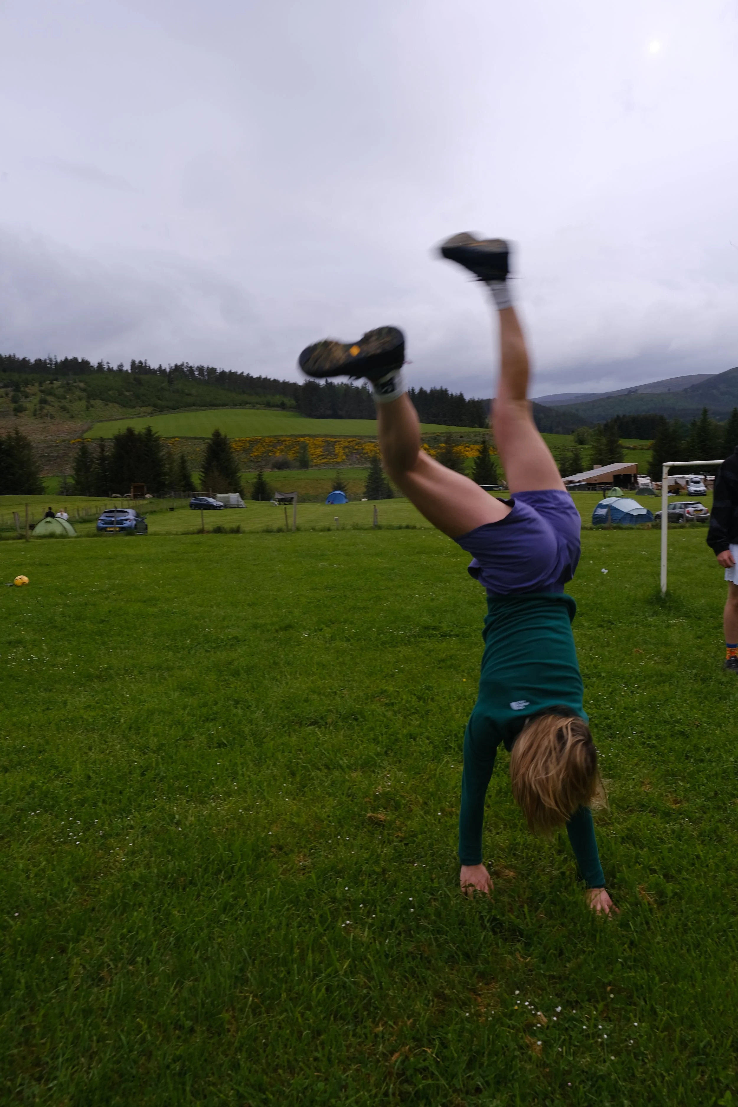 A person performing a handstand in a grassy field with hills and tents in the background.