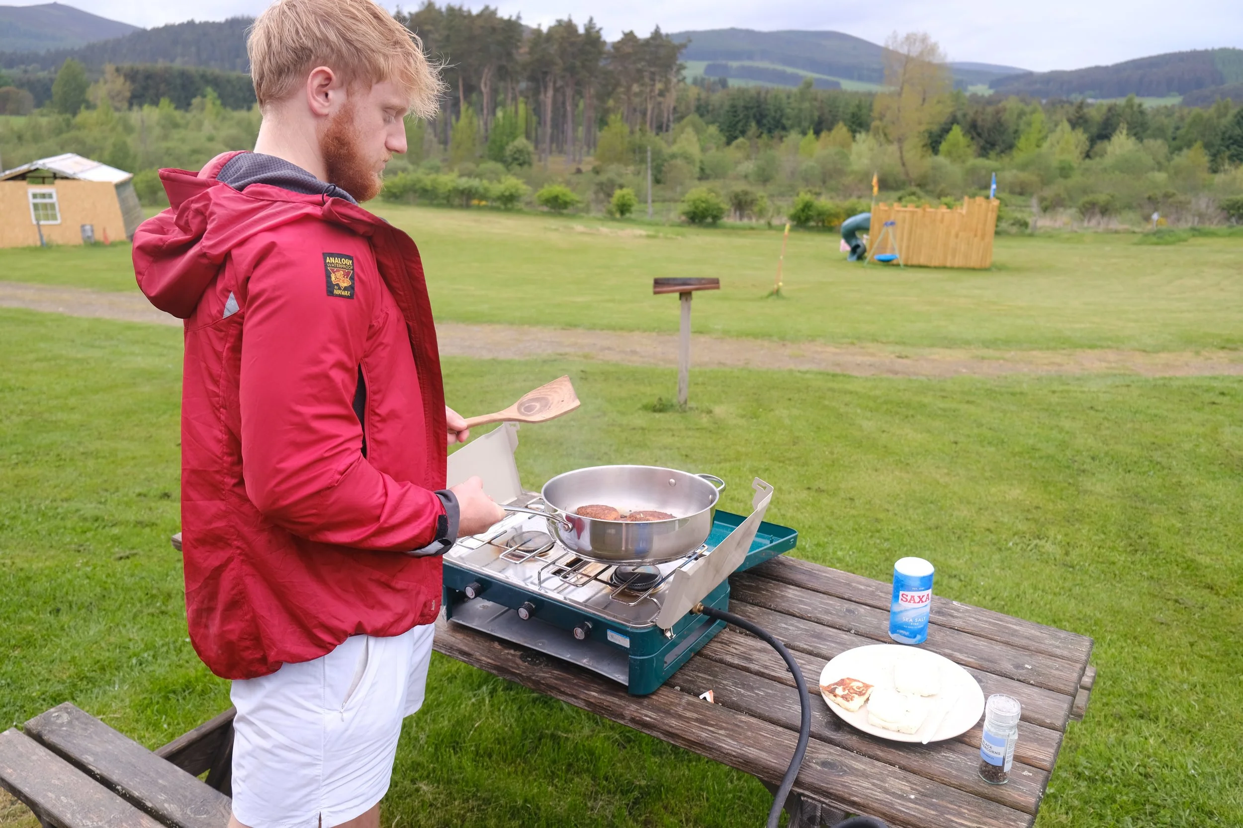 A young man with red hair and beard, wearing a red jacket and white shorts, is cooking burgers on a portable gas stove outdoors at a park. There is a wooden table with a plate of bread and condiments, and a bottle of salt on the table. The background