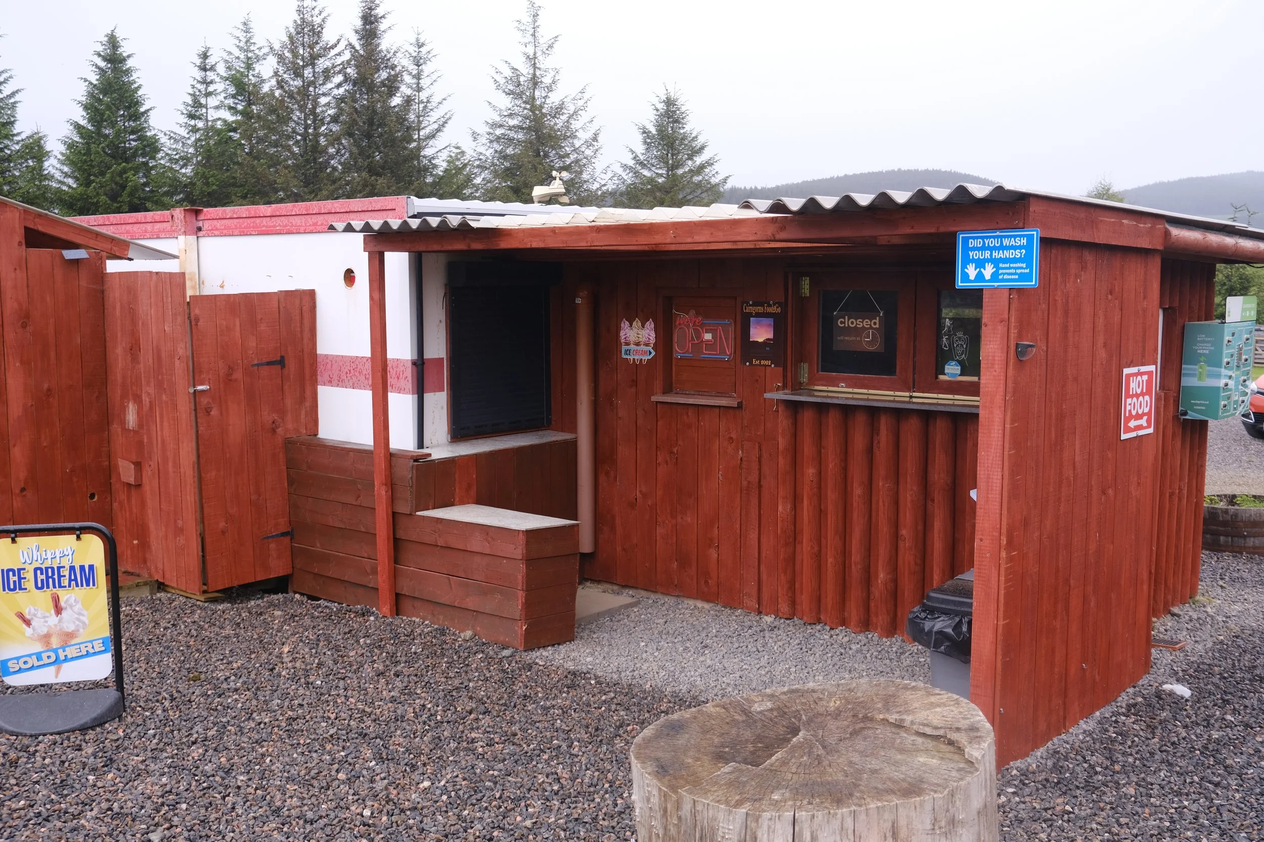Small wooden food stand with signs advertising ice cream and hot food, located outdoors on gravel with forested hills in the background.
