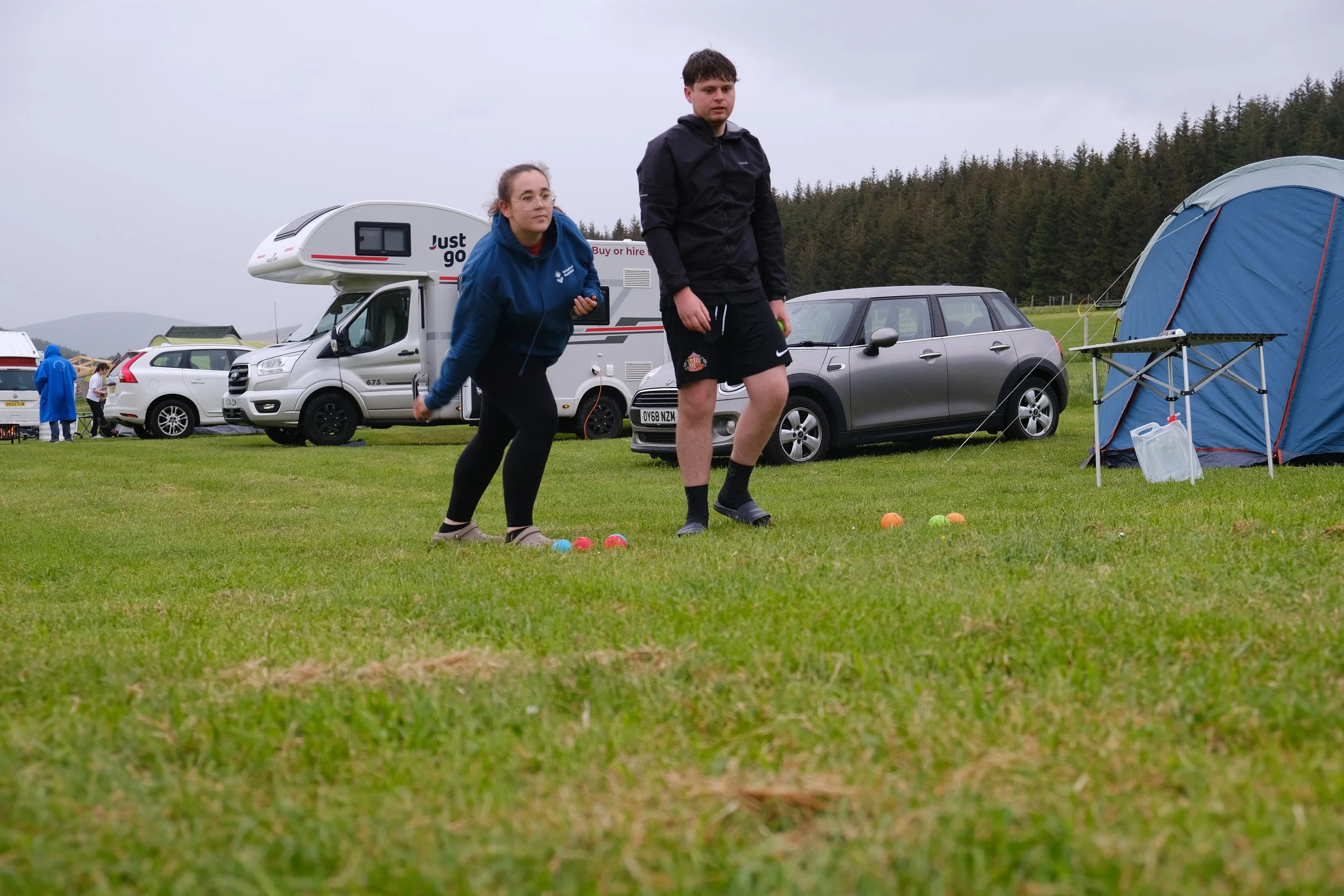 Two young people playing with colorful balls in a grassy field near tents and cars, with a wooded hillside in the background on a cloudy day.