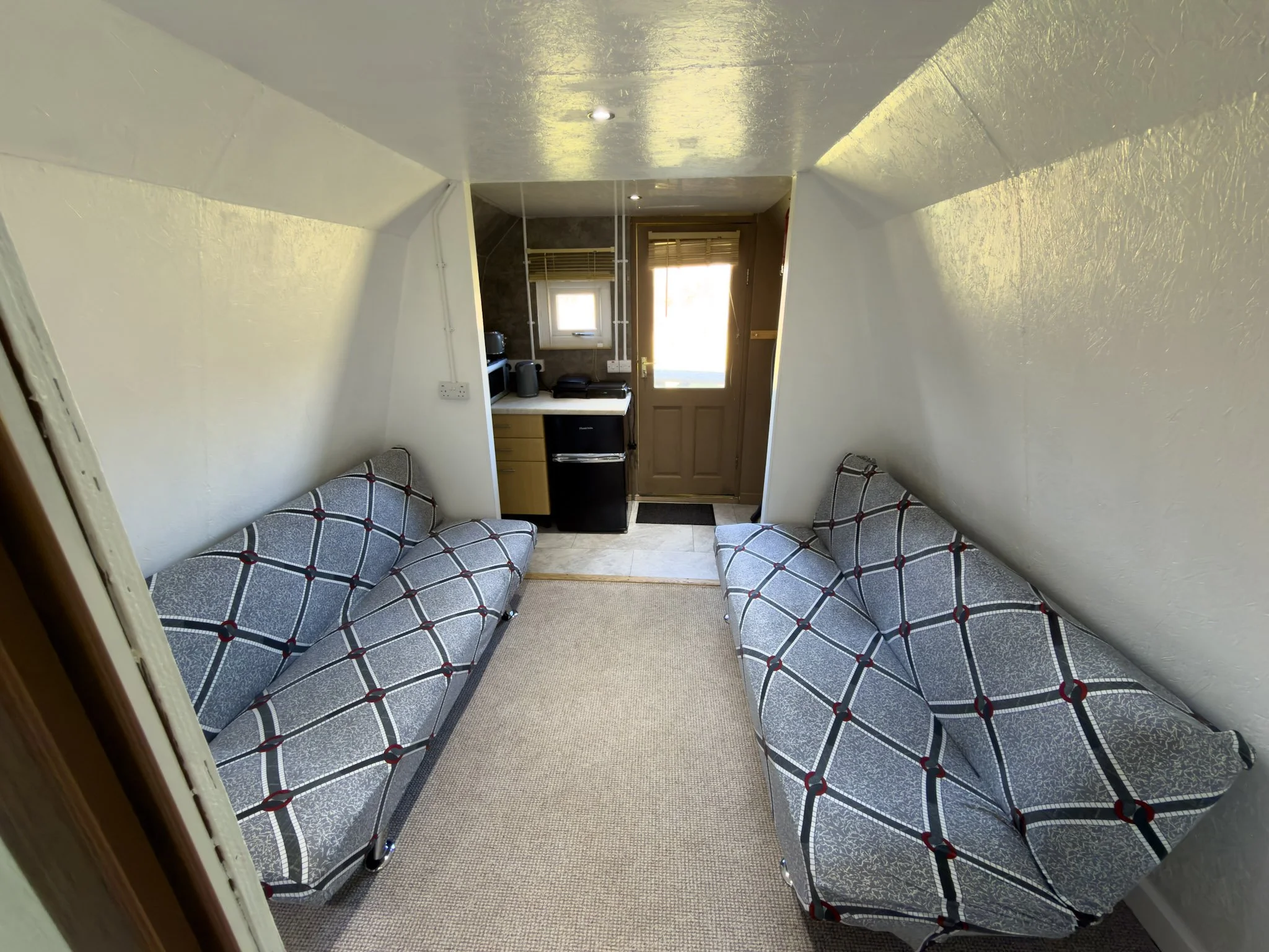 Two patterned sofas facing each other in a small, cozy room with cream carpet, a window with blinds, a door with sunlight coming through, and a small kitchenette with appliances in the background.