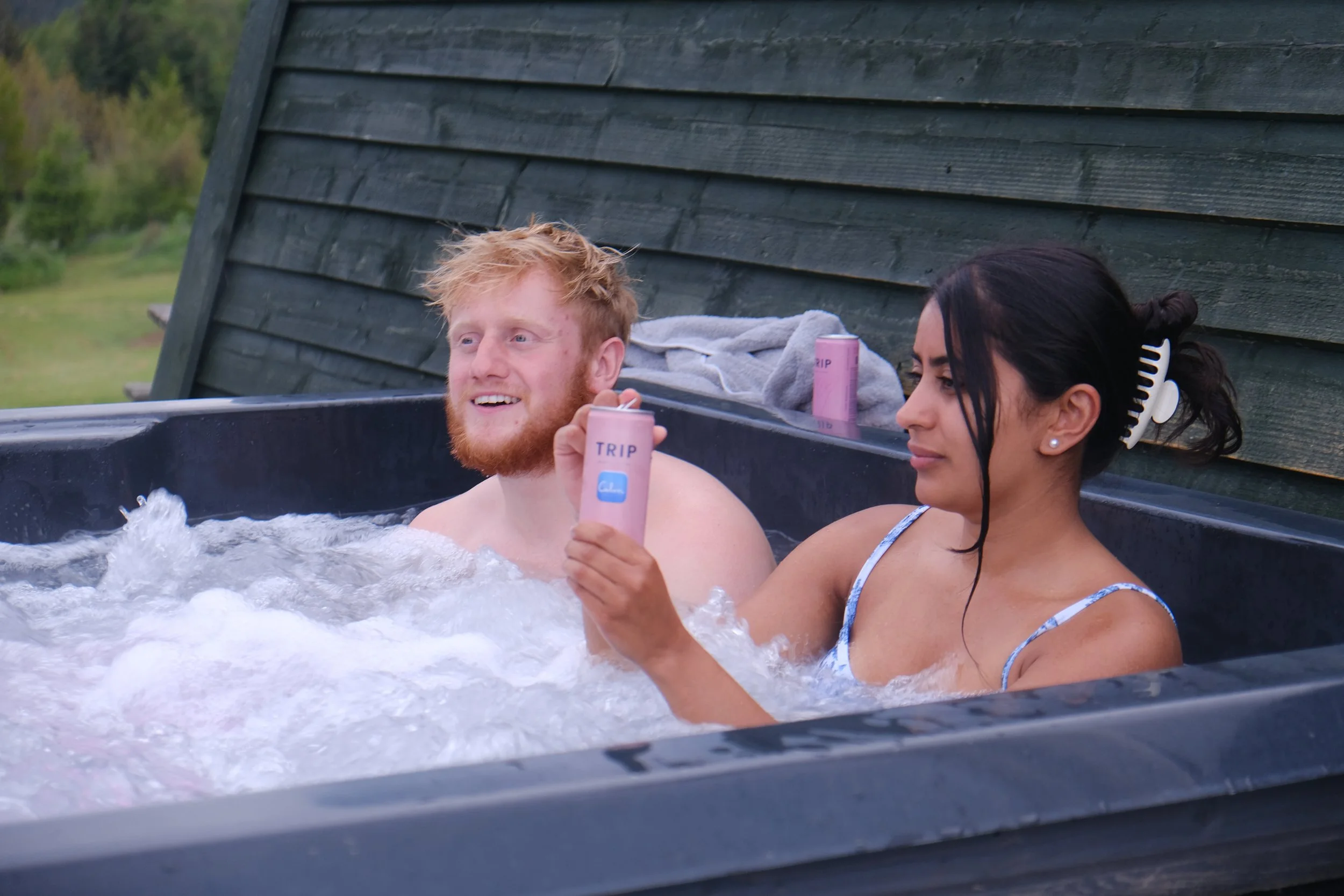 A man with red hair and beard and a woman with black hair and a white hair clip relaxing in a hot tub outdoors, each holding a can of Tripping Pink beverage.