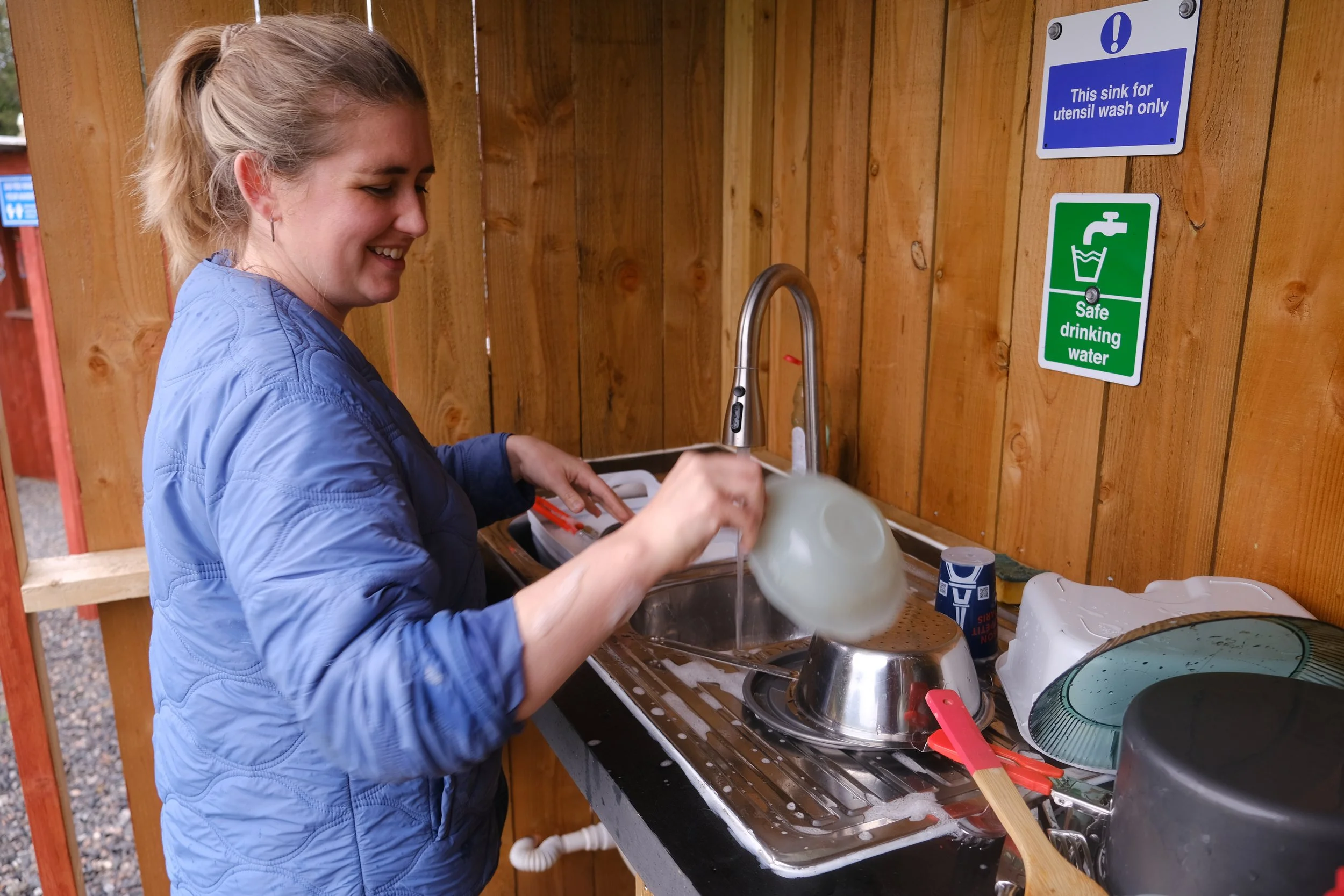 A woman washing dishes at an outdoor sink with a wooden backsplash, wearing a blue jacket and smiling.
